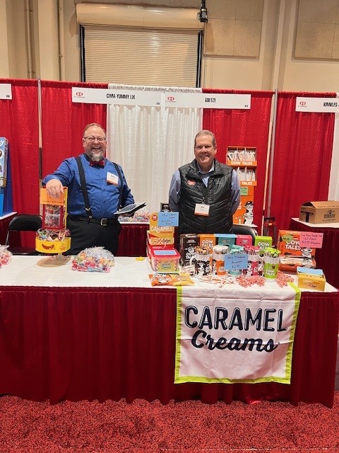 Two men, one in a bow tie and suspenders, smile behind a table with a Caramel Creams cloth and Cow Tales boxes.