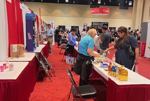 Attendees browsing candy on red-skirted tables in a convention hall aisle.