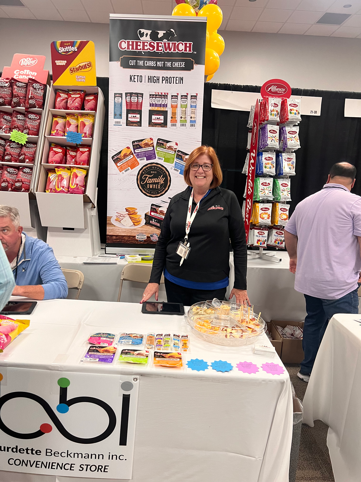 Woman with a lanyard stands behind a bbi Convenience Store table in front of a Cheesewich banner and candy displays.