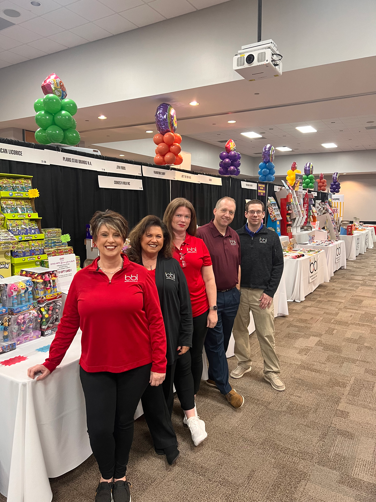 Five people in red and black bbi shirts pose beside white-draped booths under balloon columns in a trade show hall.