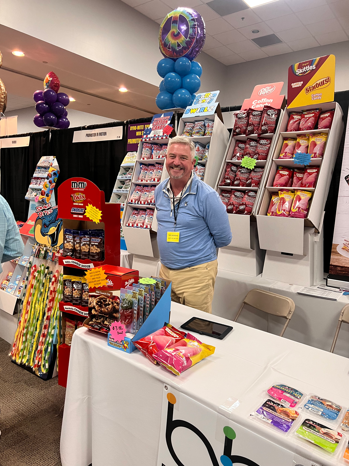 Smiling man in a blue pullover stands beside Dr Pepper and Skittles cotton candy displays behind a white table.