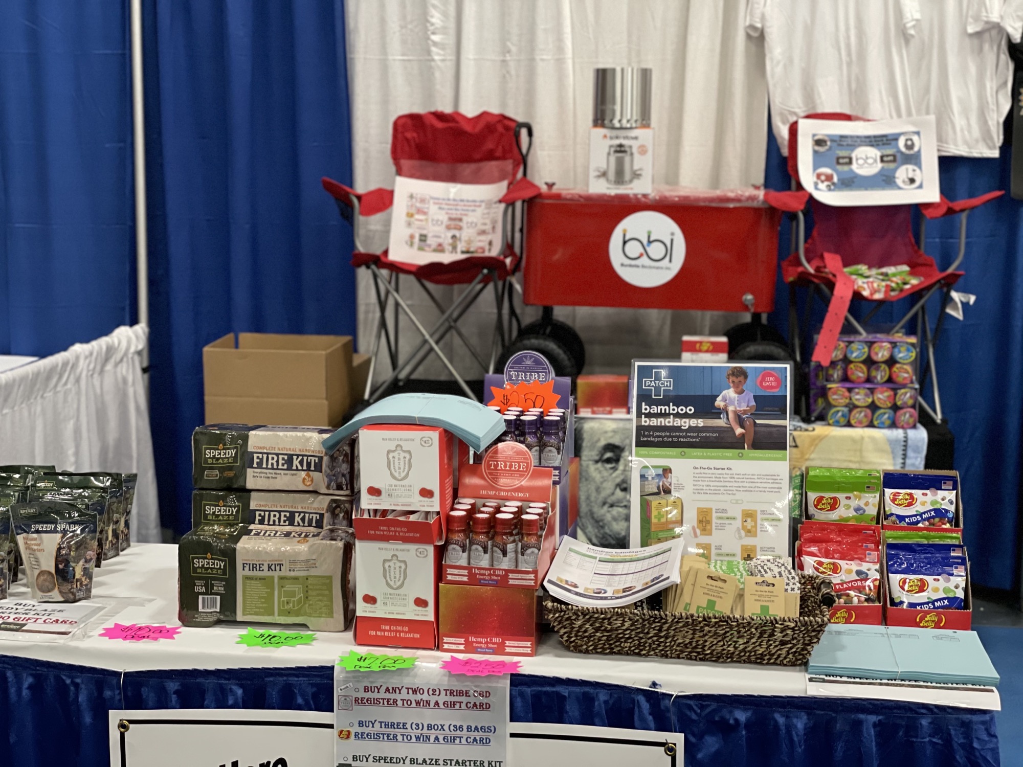 Booth table with fire kits, bottles and bamboo bandages in front of a red bbi wagon cooler and red chairs.