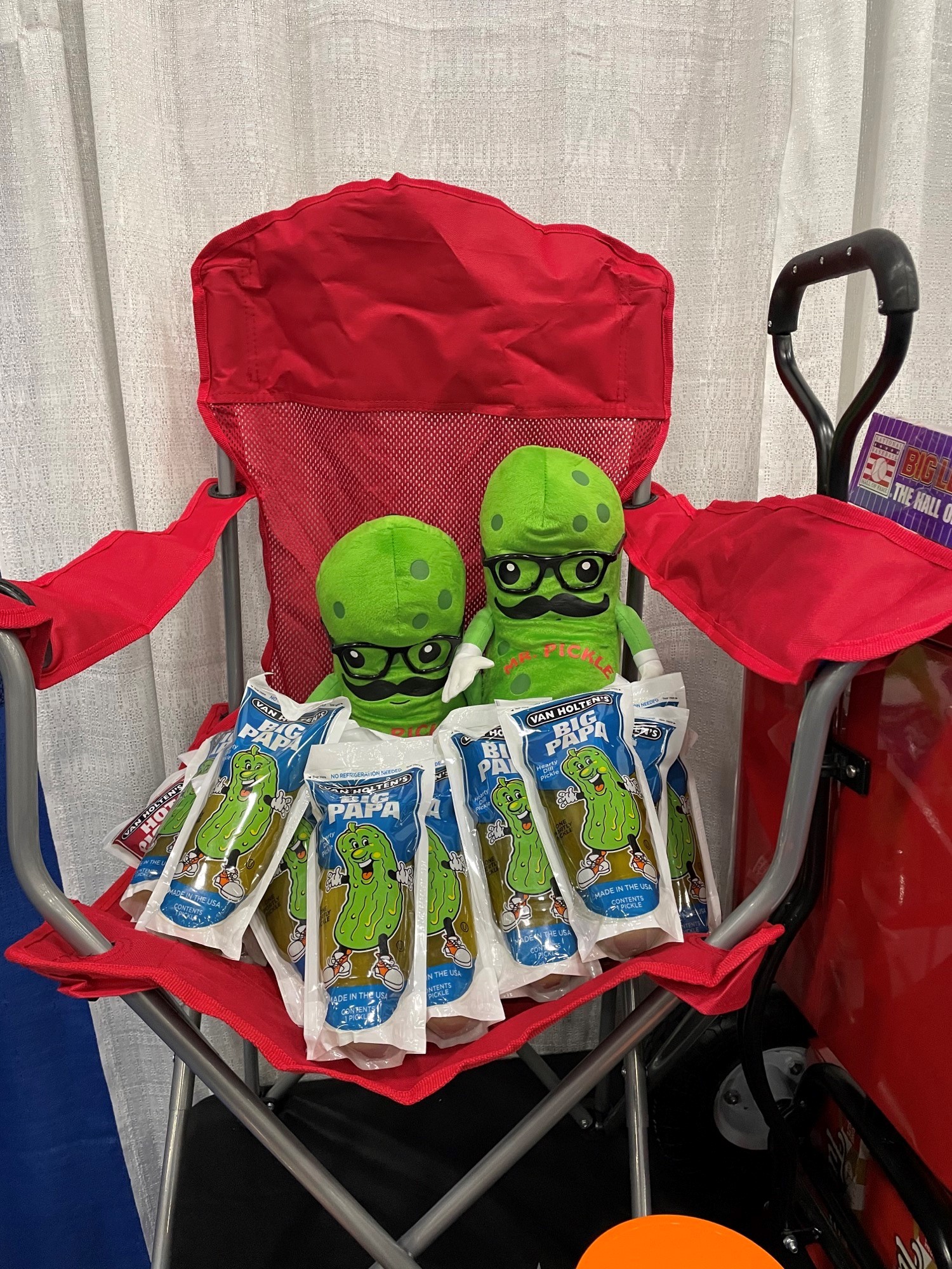 Two green plush pickles in glasses and mustaches sit on a red chair surrounded by Van Holten's Big Papa pickle pouches.