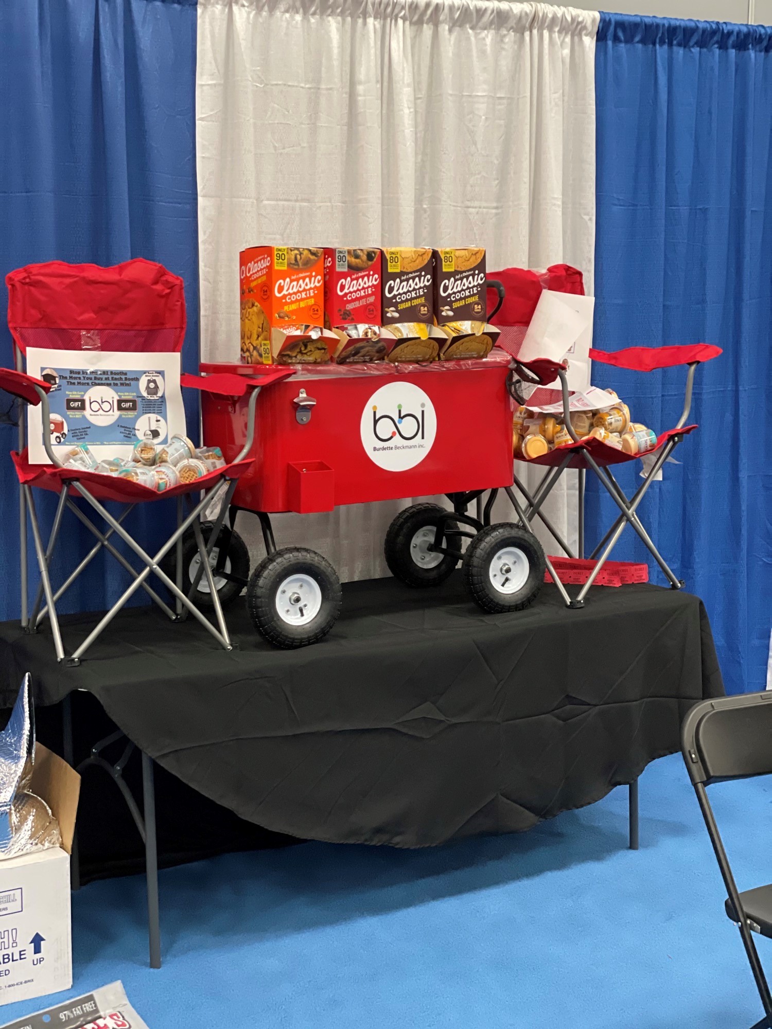 Red bbi wagon cooler topped with Classic Cookie boxes sits on a black-draped table between red chairs holding small items.