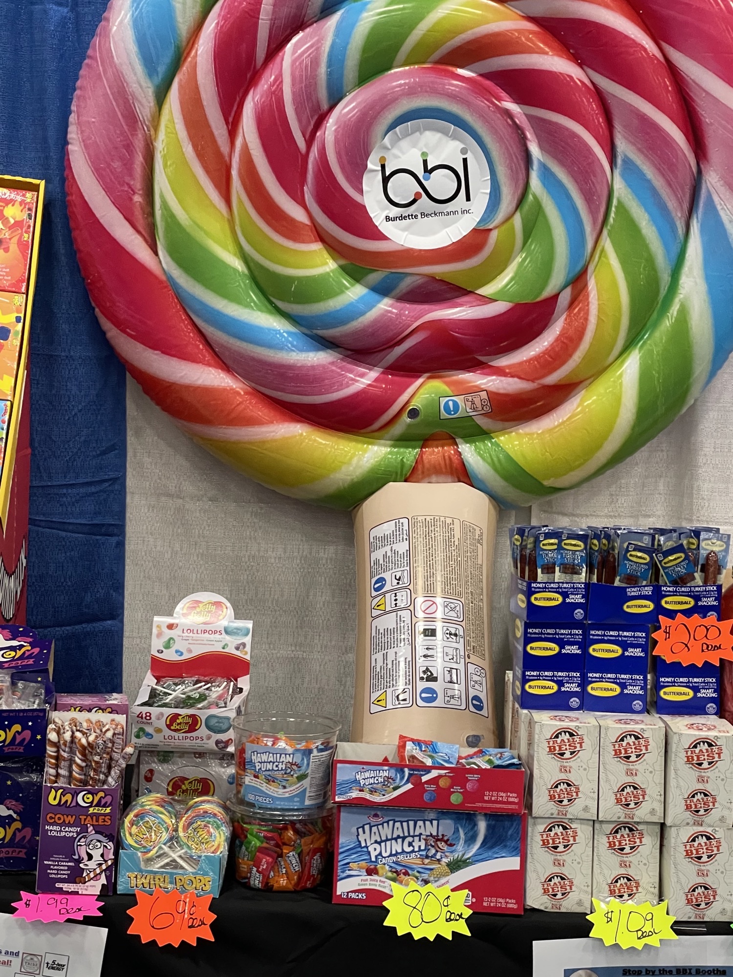 Giant inflatable rainbow lollipop with a bbi logo above candy, Hawaiian Punch and beef stick displays with price tags.