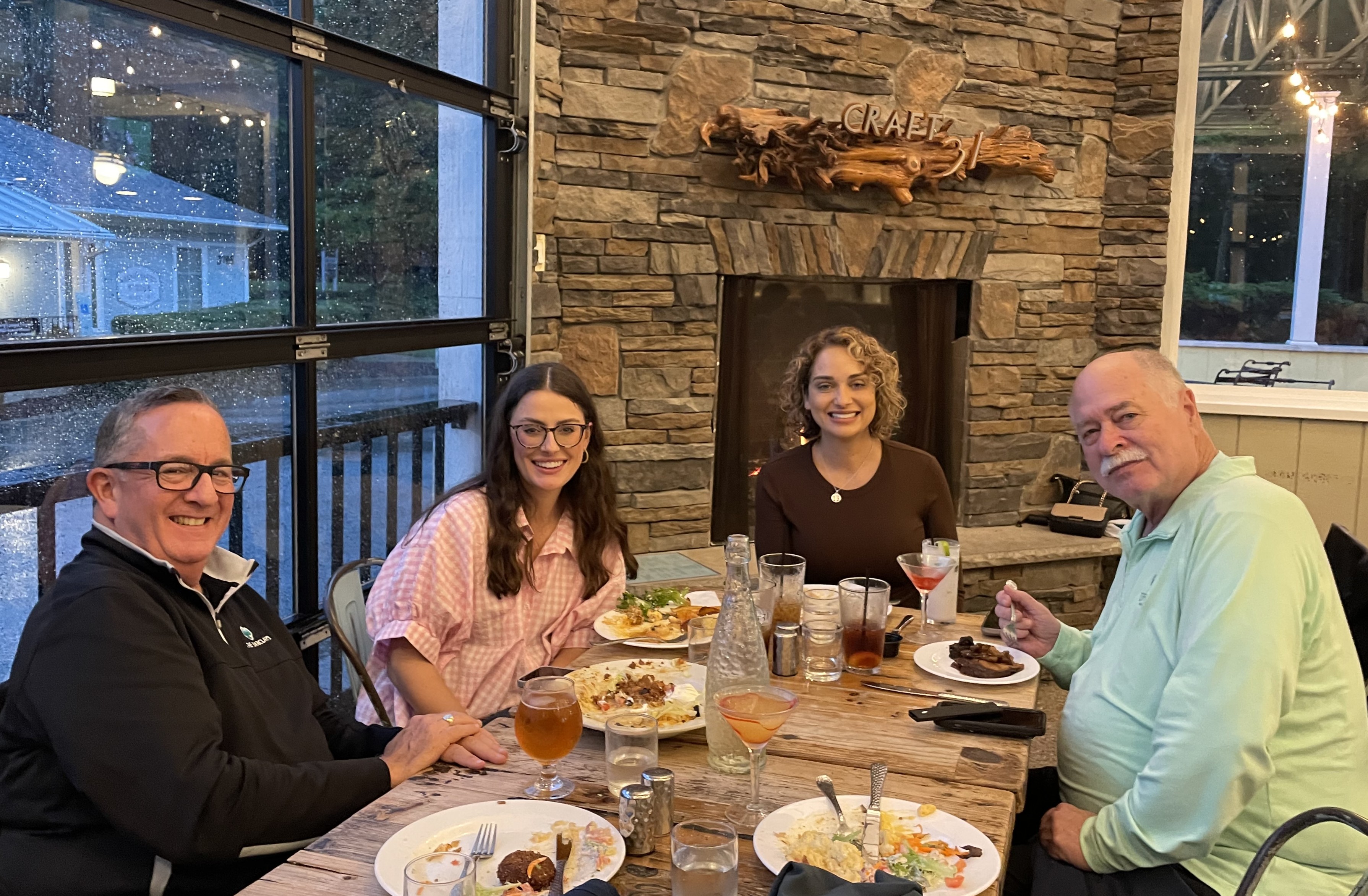 Four people share a meal at a rustic wooden table in front of a stone fireplace.