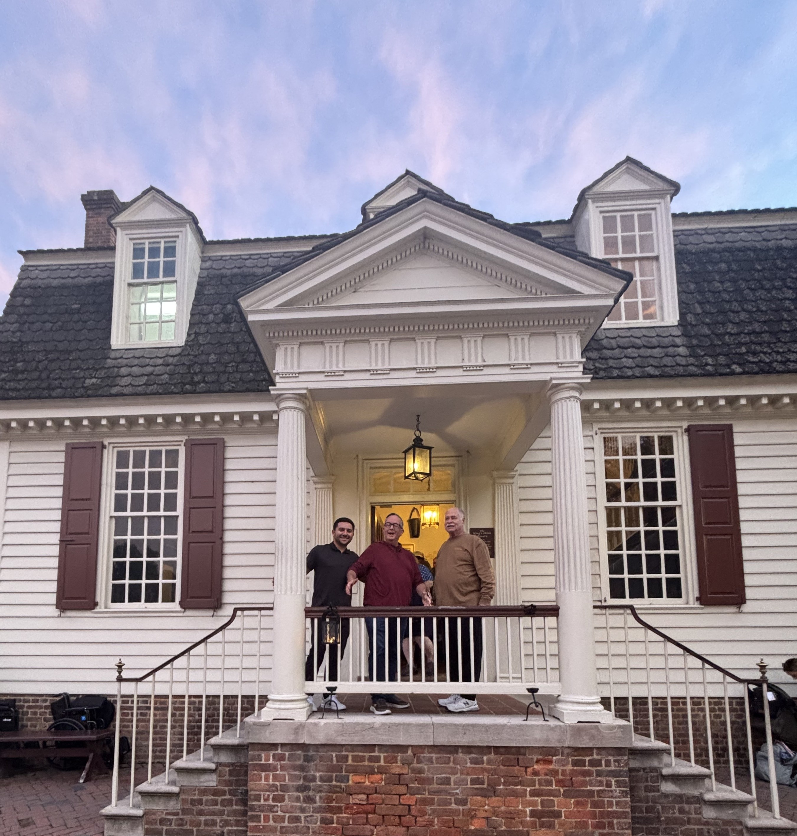 Three men stand on the porch of a white colonial building with columns at dusk.