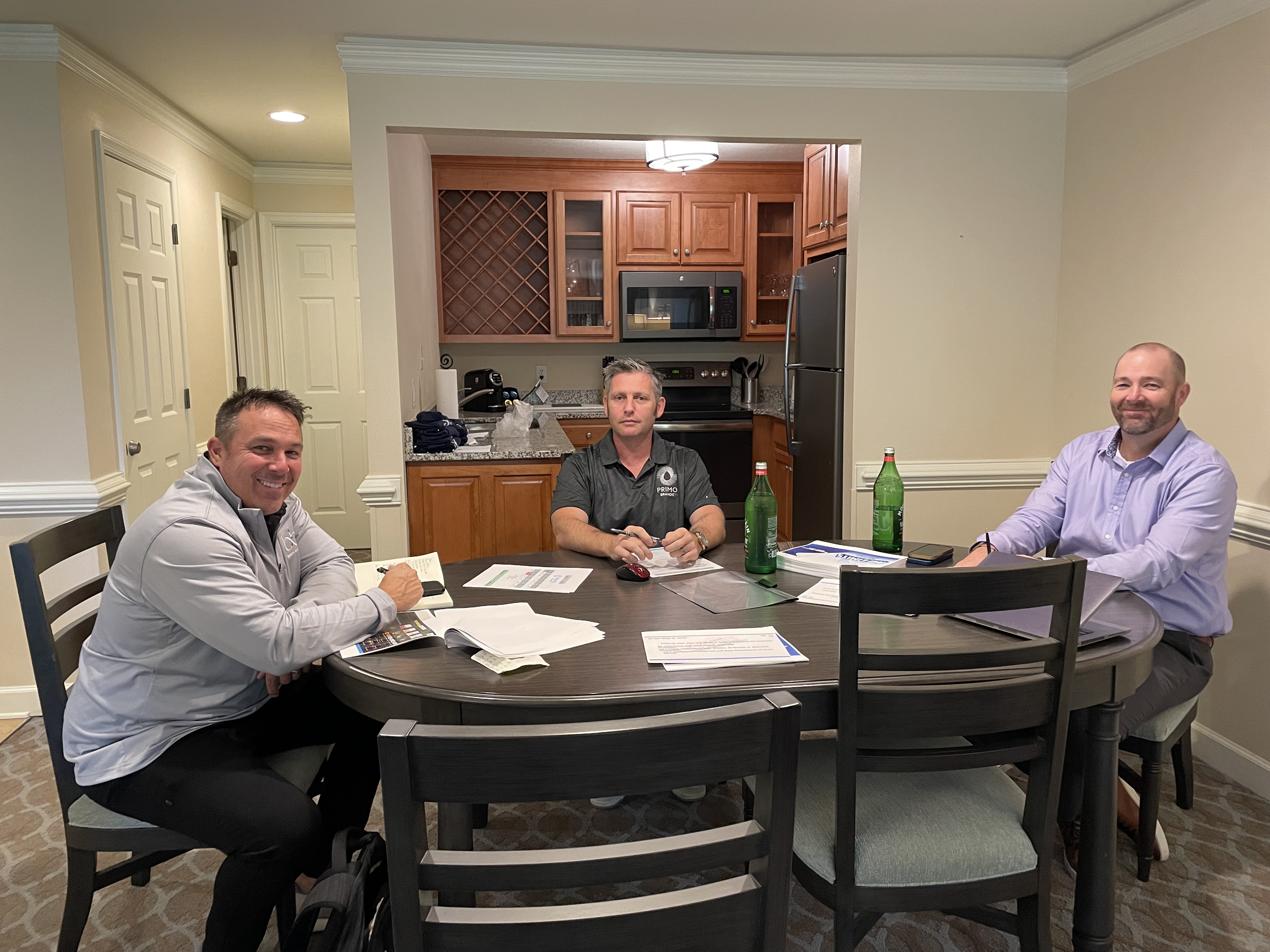 Three men sit at a round table in a hotel suite with printed papers and green bottles.
