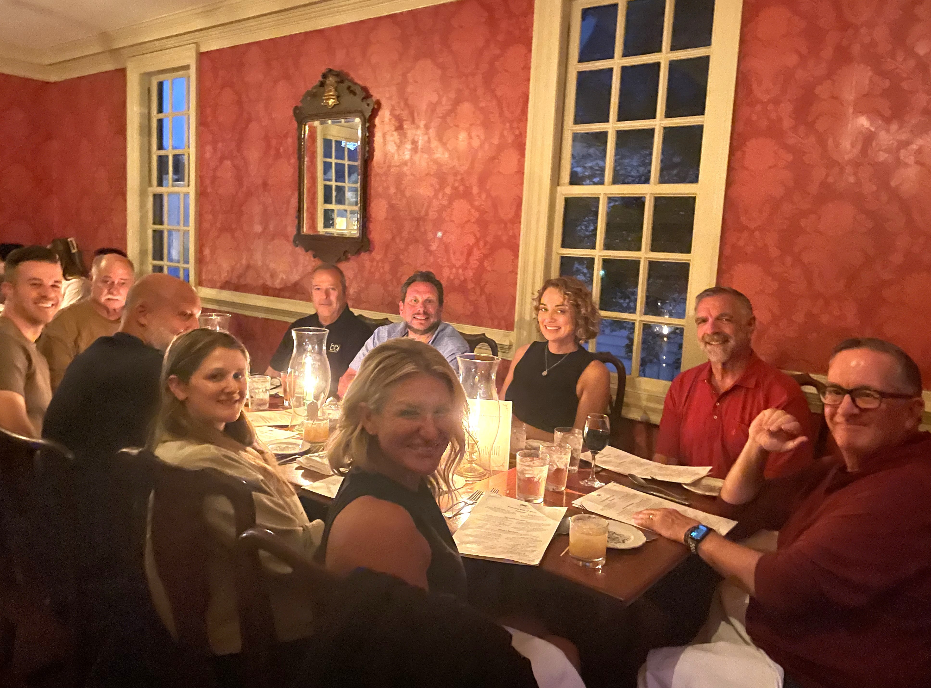 Group dinner in a dim, candlelit, red-walled colonial dining room, with diners leaning toward the camera.