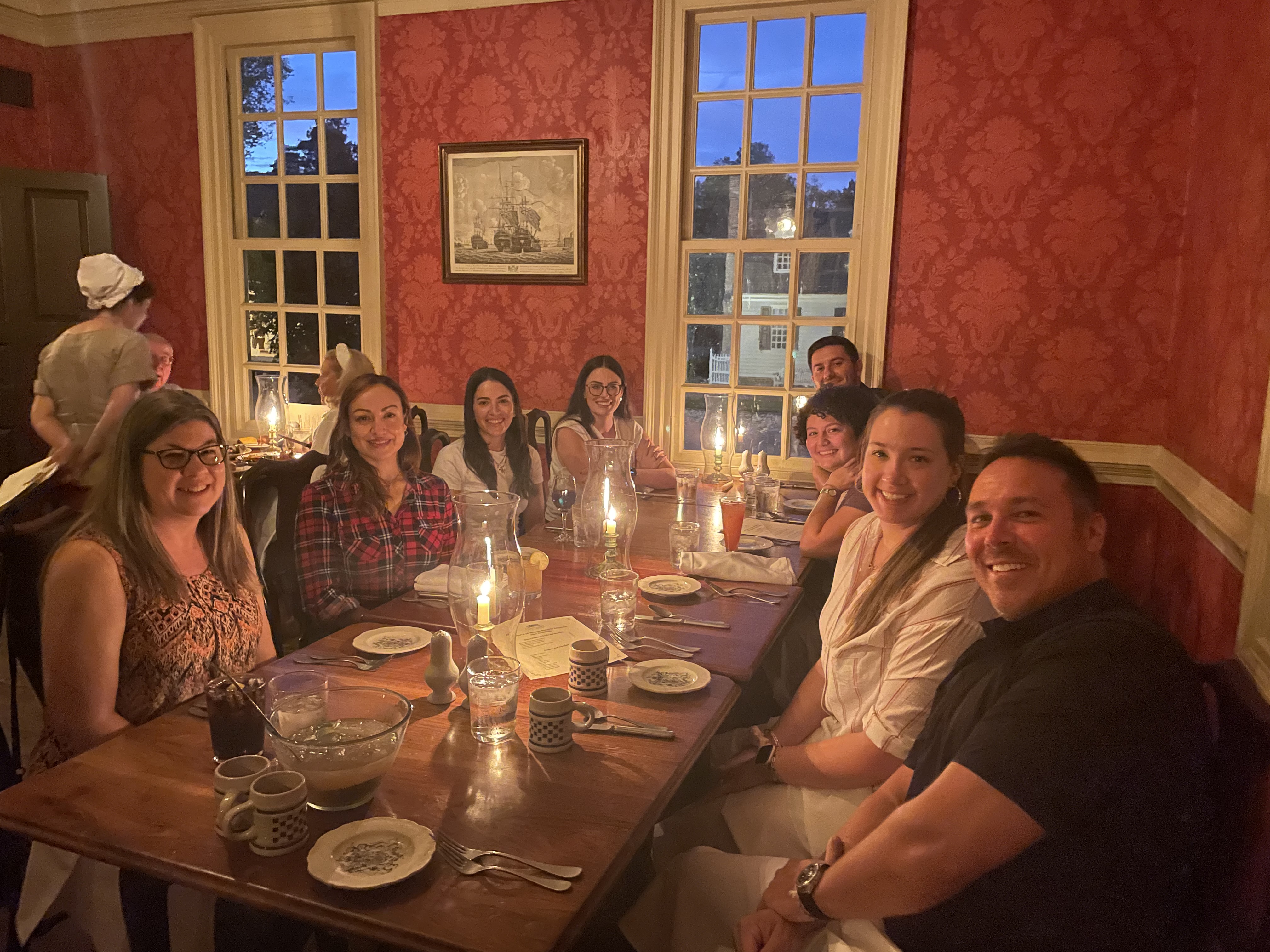 Group of adults smile around a candlelit table in a red-wallpapered colonial dining room.
