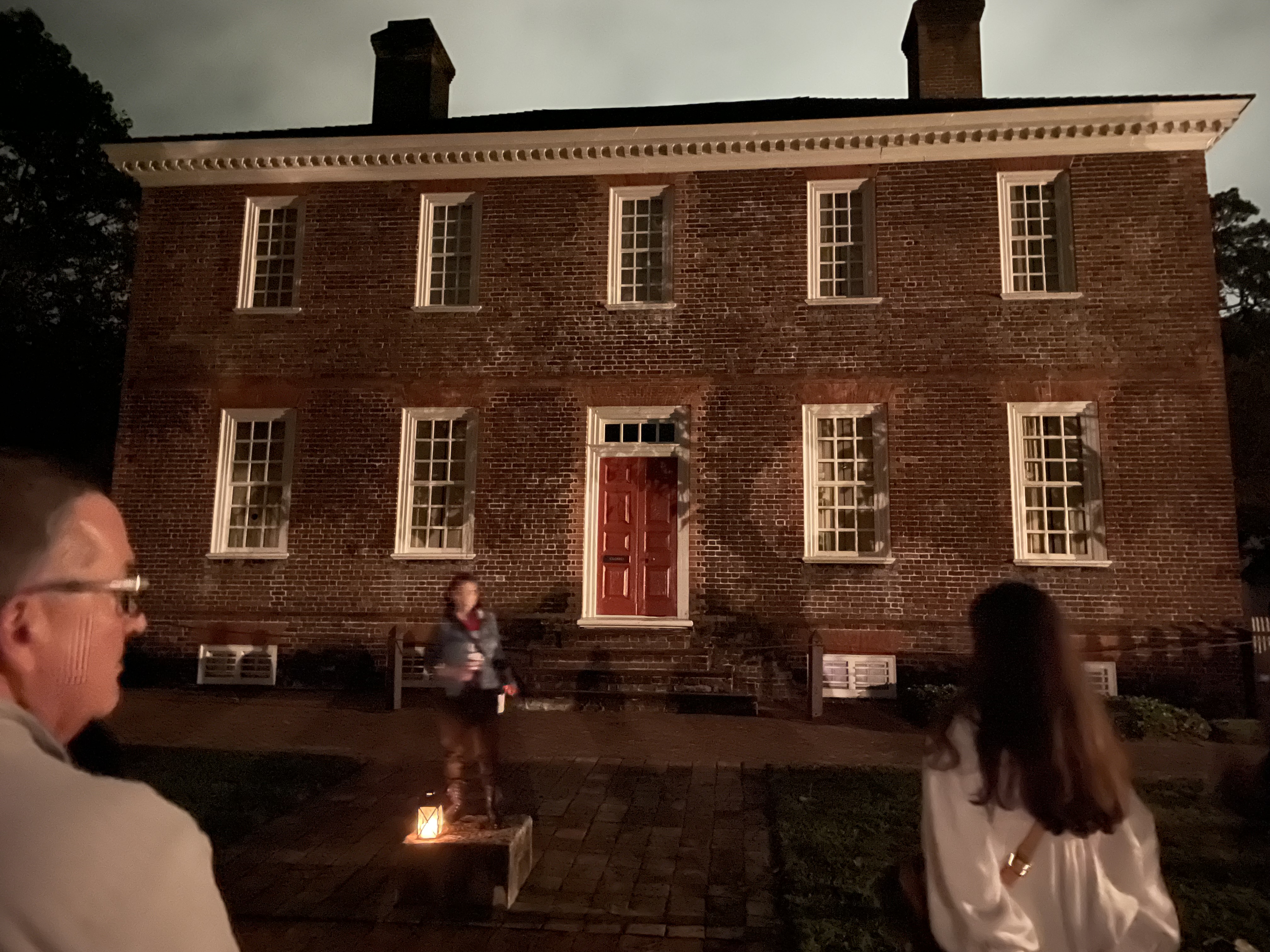 Brick colonial building at night with a red door, a person standing in front and two spectators in the foreground.