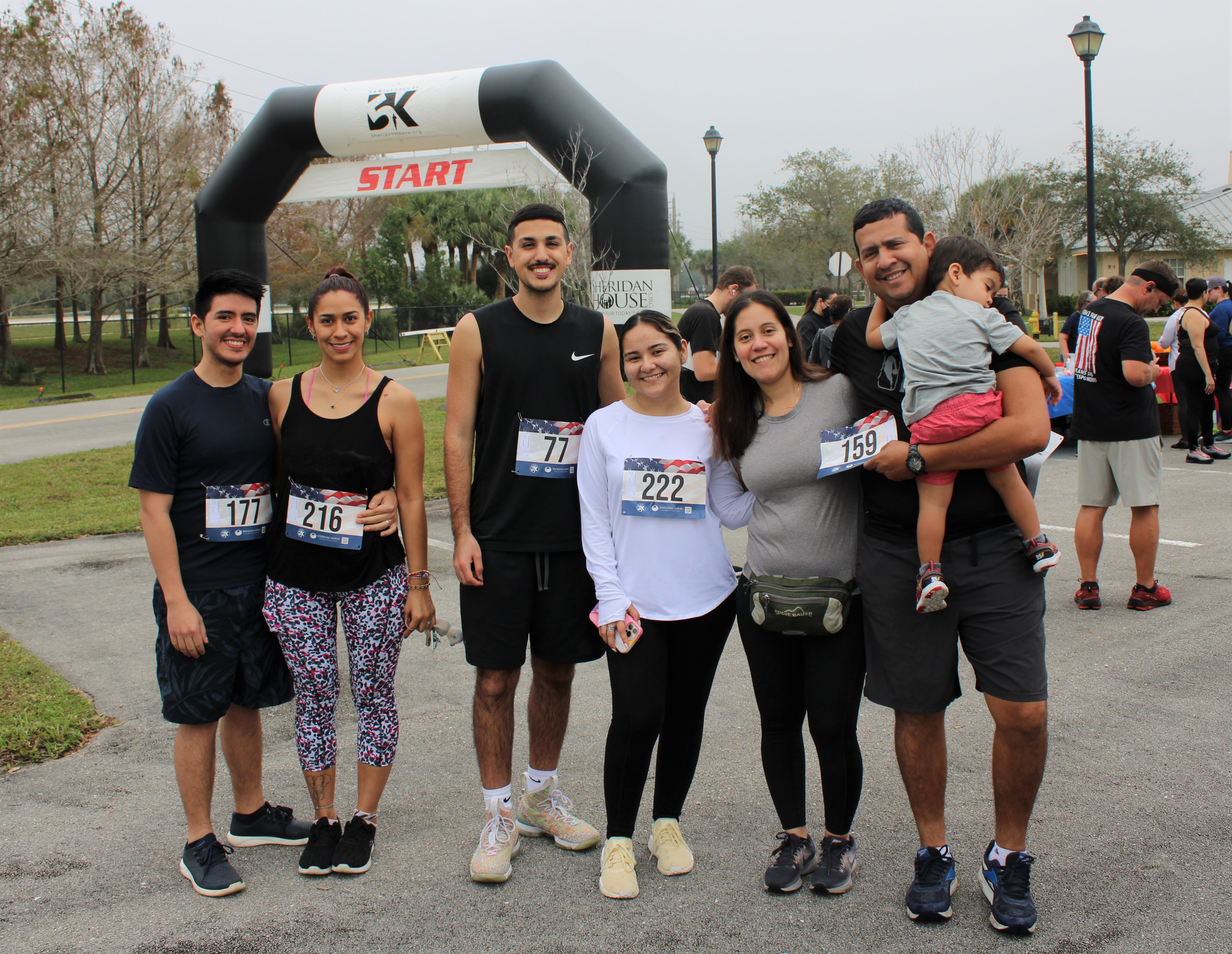 Six smiling participants, most wearing race bibs, pose in a row, one man holding a toddler, with the START arch behind.