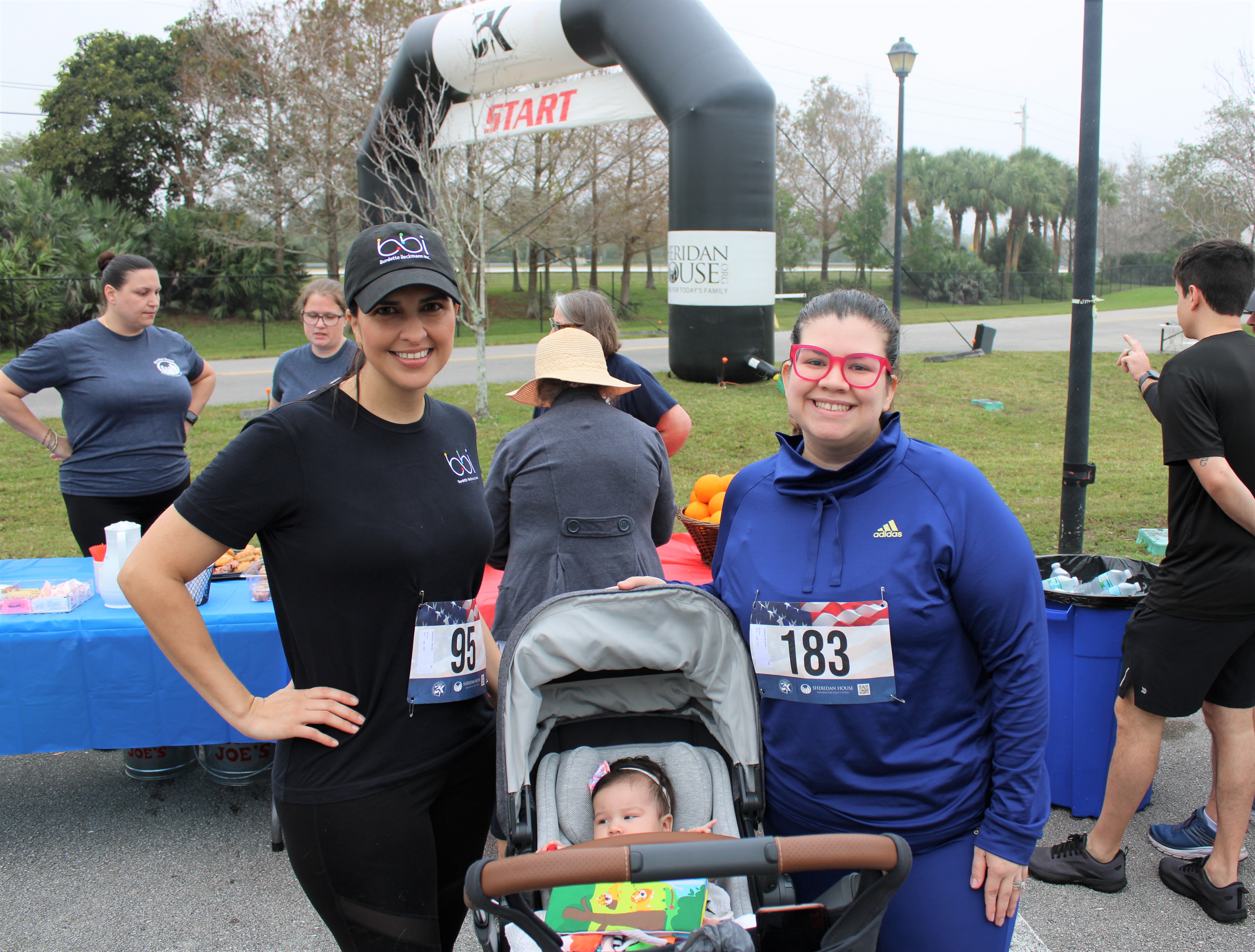 A woman in a bbi cap and a woman in red glasses and a blue pullover pose beside a stroller with a baby at the start.