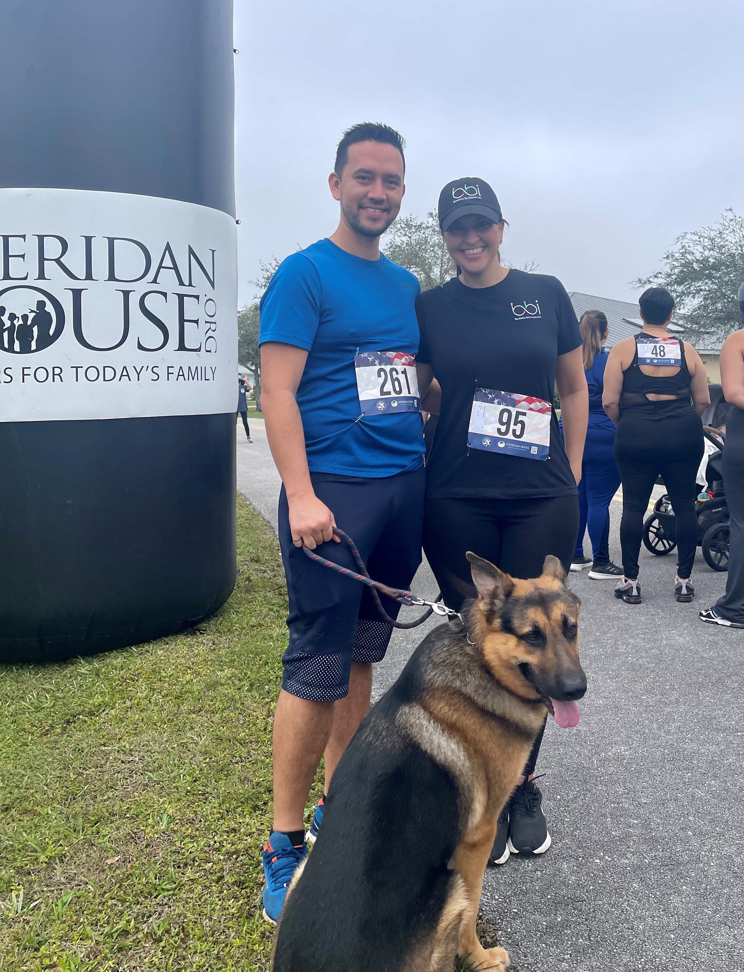 A man with a leashed German shepherd and a woman in a bbi cap pose beside a Sheridan House inflatable column.