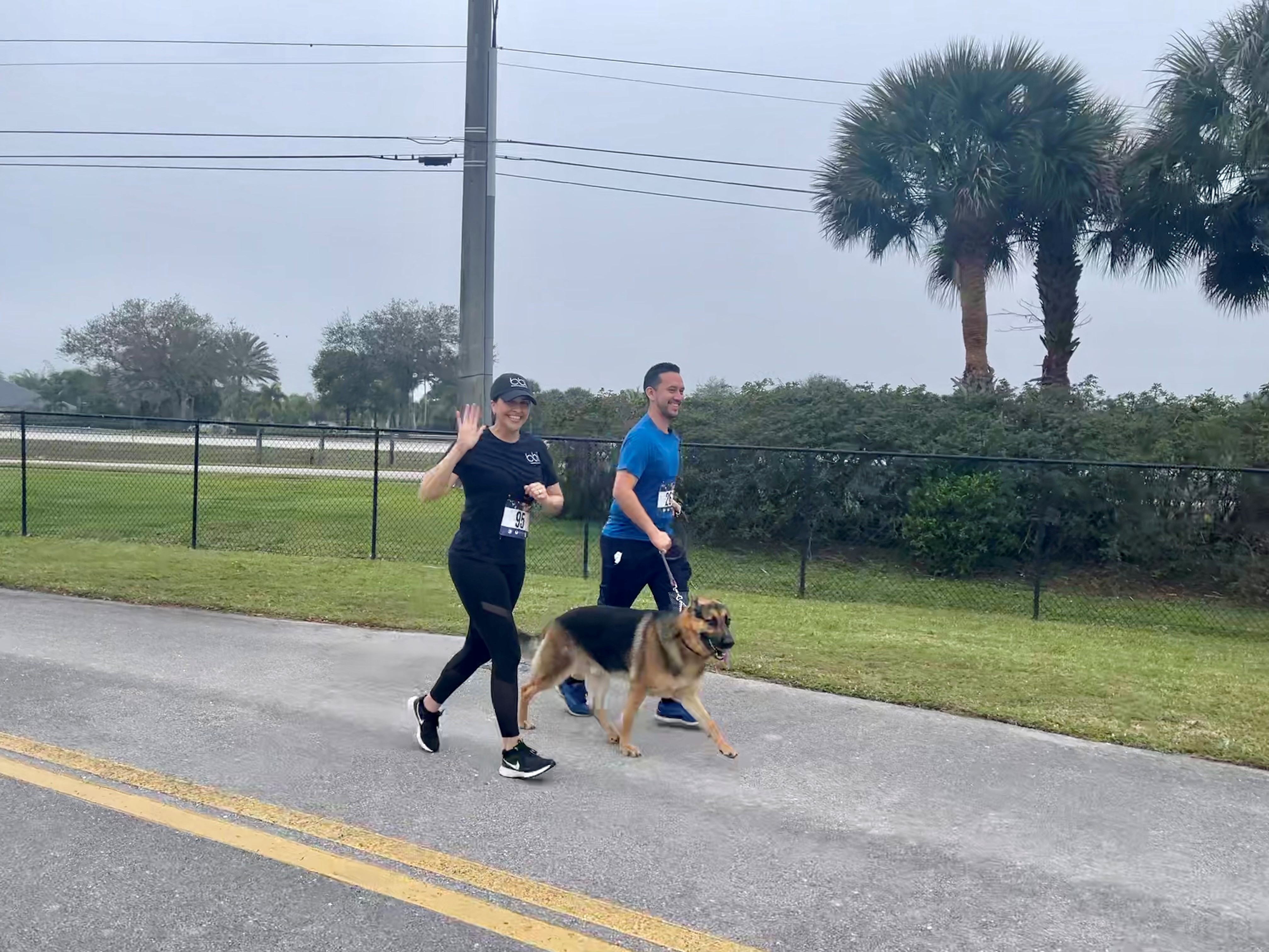 A woman in a cap waves as she and a man in blue walk a leashed German shepherd along a paved path with palm trees behind.