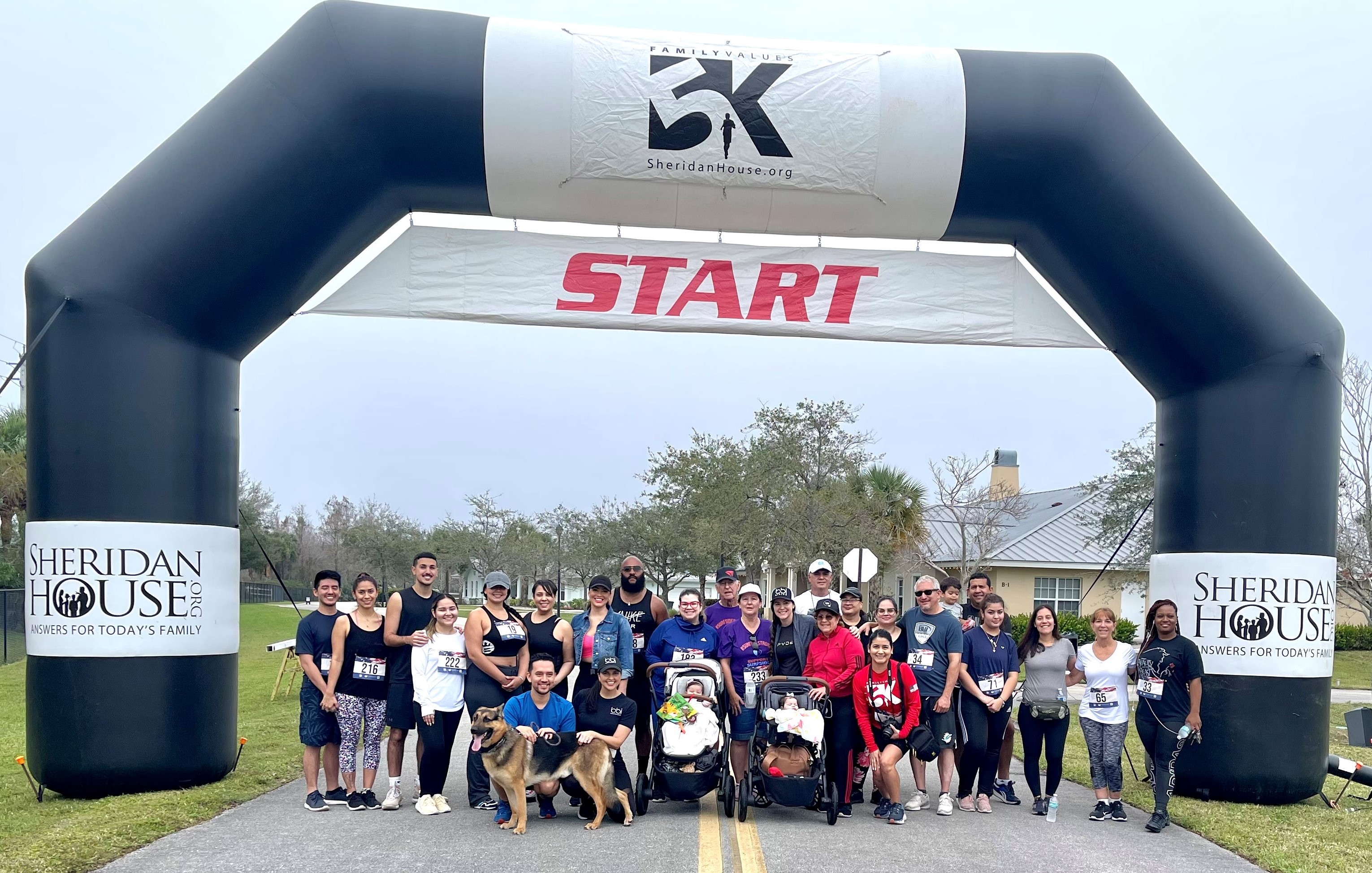 Runners, walkers, strollers and a German shepherd gather under a black inflatable START arch for the Sheridan House 5K.