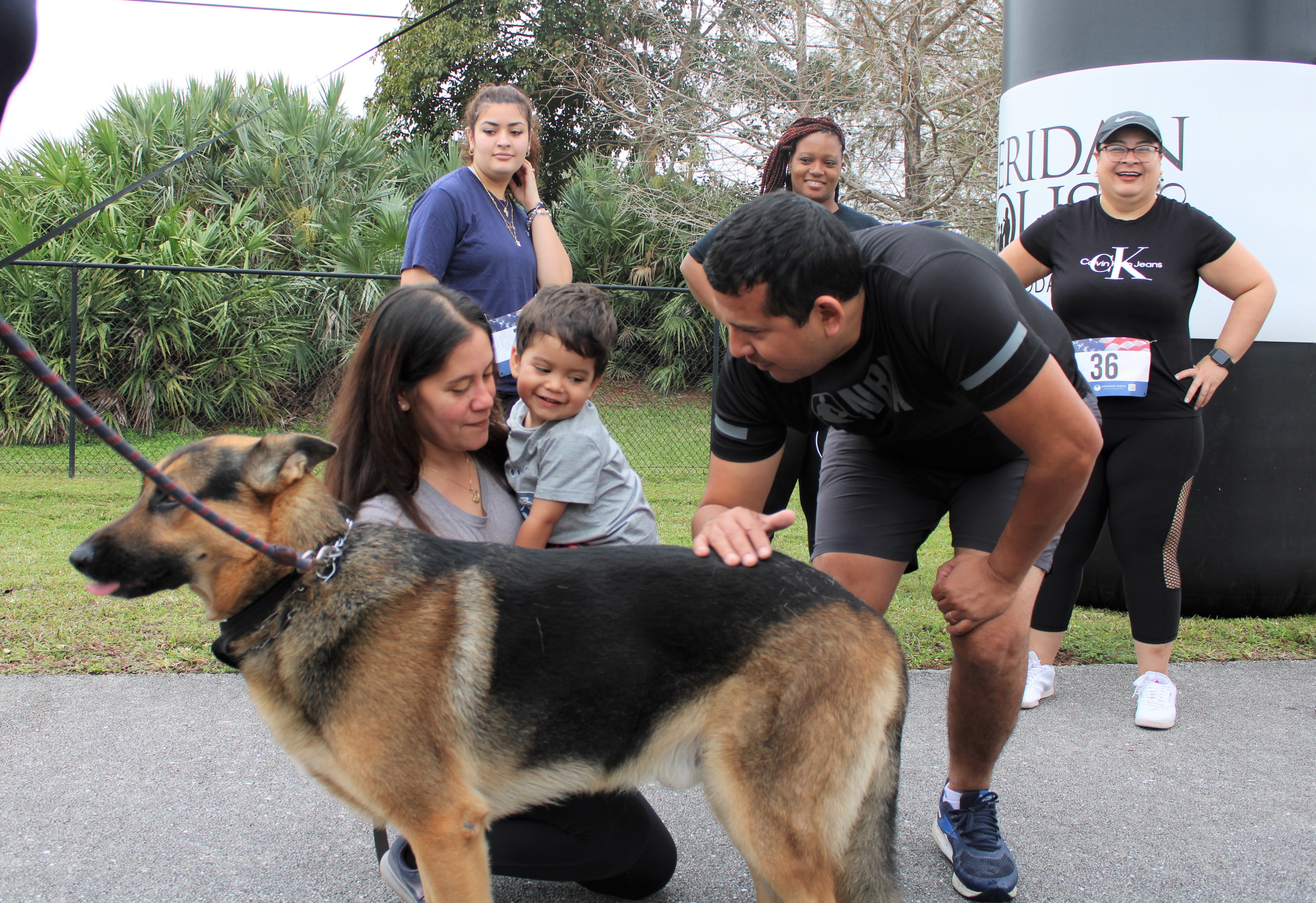 A woman kneels holding a toddler while a man bends to pet a German shepherd near the Sheridan House inflatable.