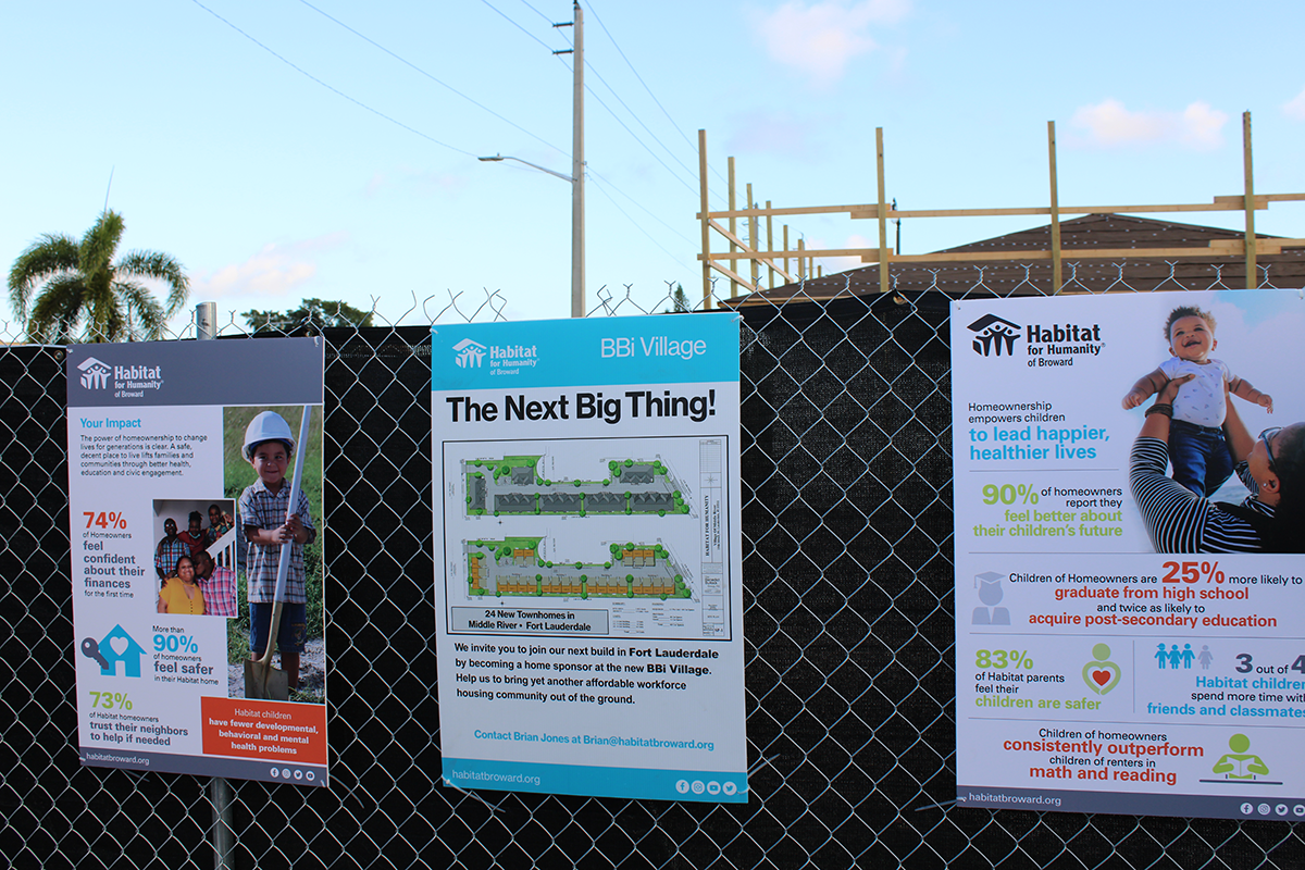 Three Habitat for Humanity posters on a chain-link fence, including one reading BBi Village The Next Big Thing.