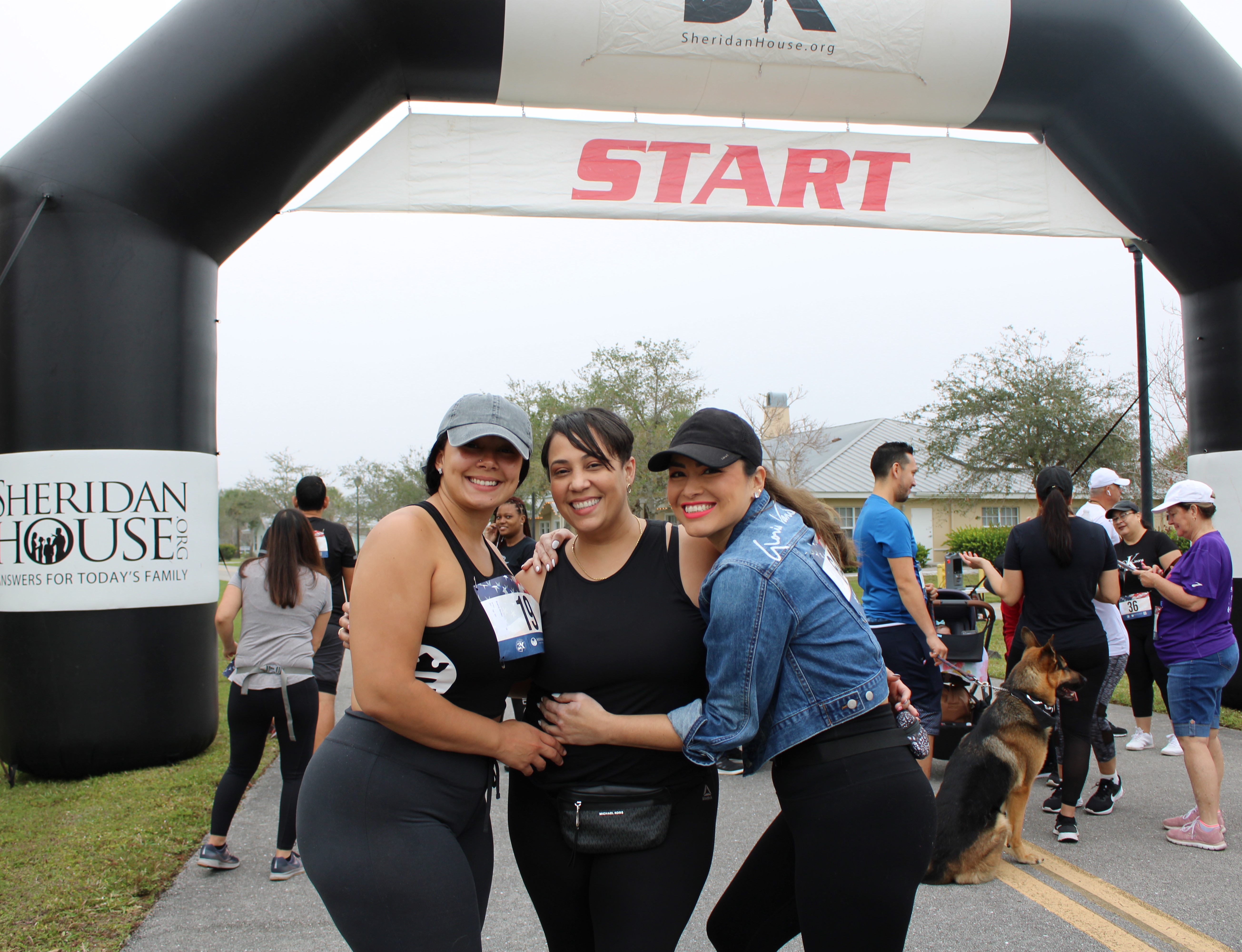 Three women hug and smile in front of the START arch, with other participants and a dog behind them.