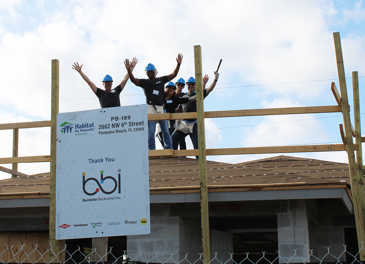 Volunteers in hard hats wave from a roof frame above a sign thanking bbi for a Pompano Beach Habitat home.