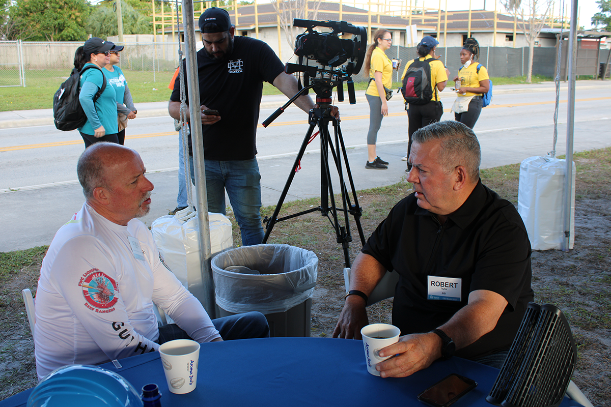 Two men talk over coffee at a blue table under a tent, with a video camera and operator behind them.