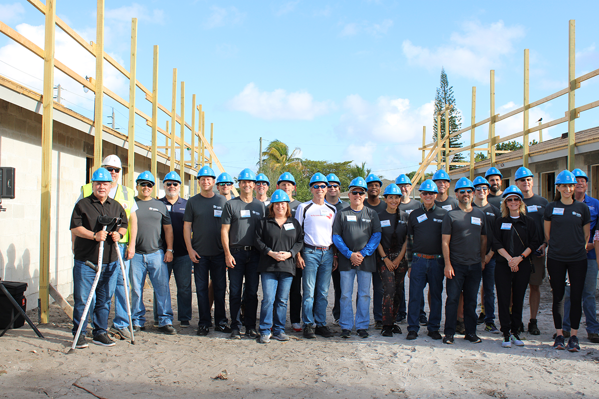 About thirty volunteers in dark shirts and blue hard hats pose on sand in front of a house frame with wood scaffolding.