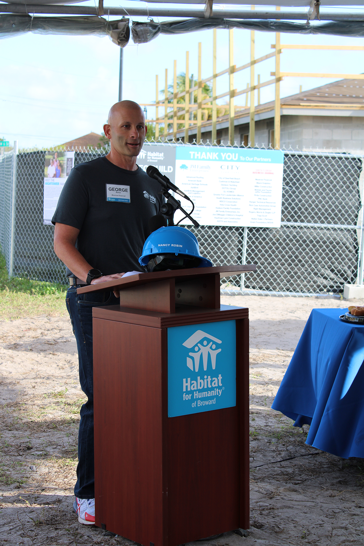 A man with a name badge speaks at a Habitat for Humanity of Broward podium, with a blue hard hat beside him.