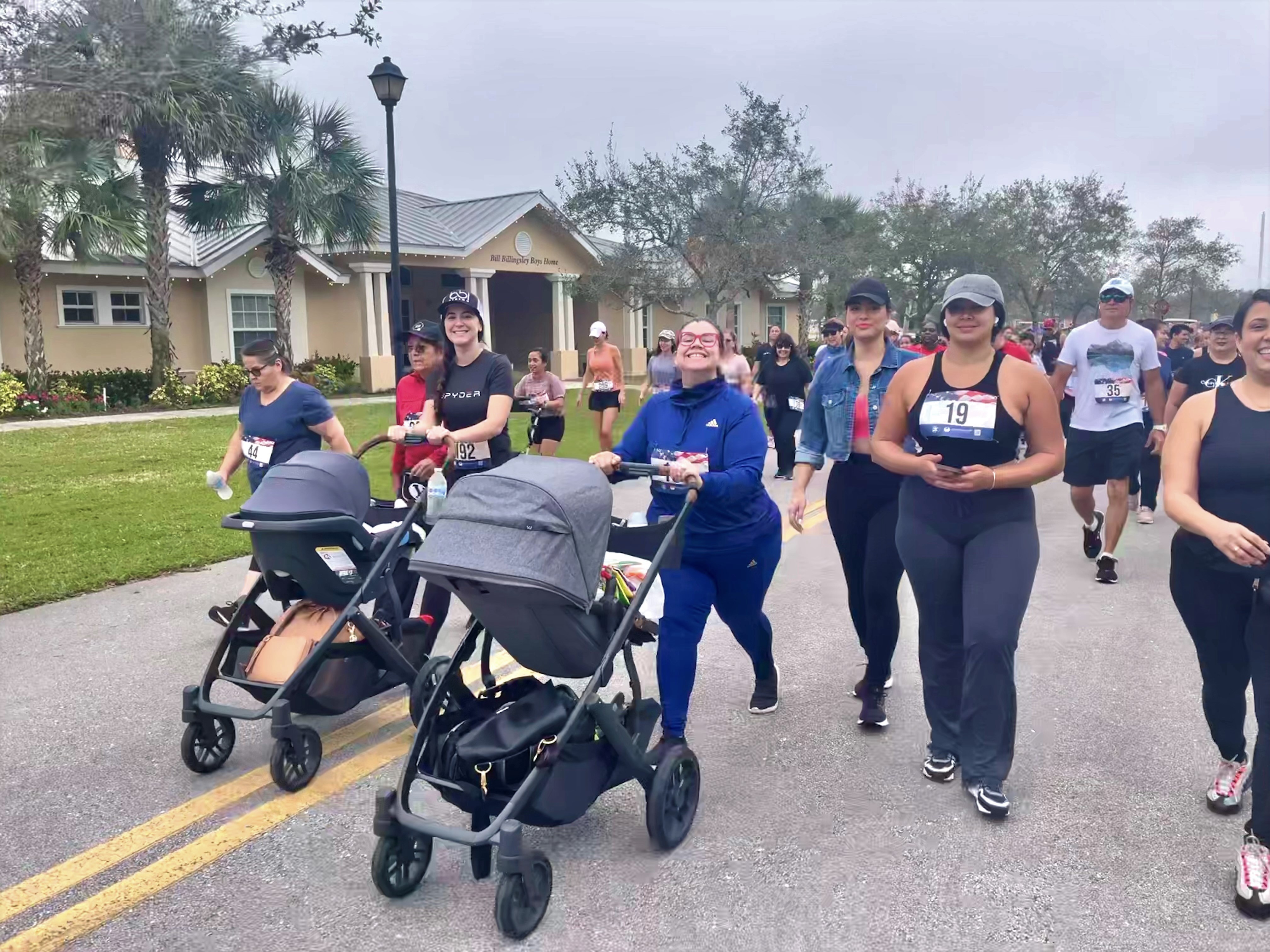 Walkers and two women pushing strollers move along a paved path past a low building at the 5K.