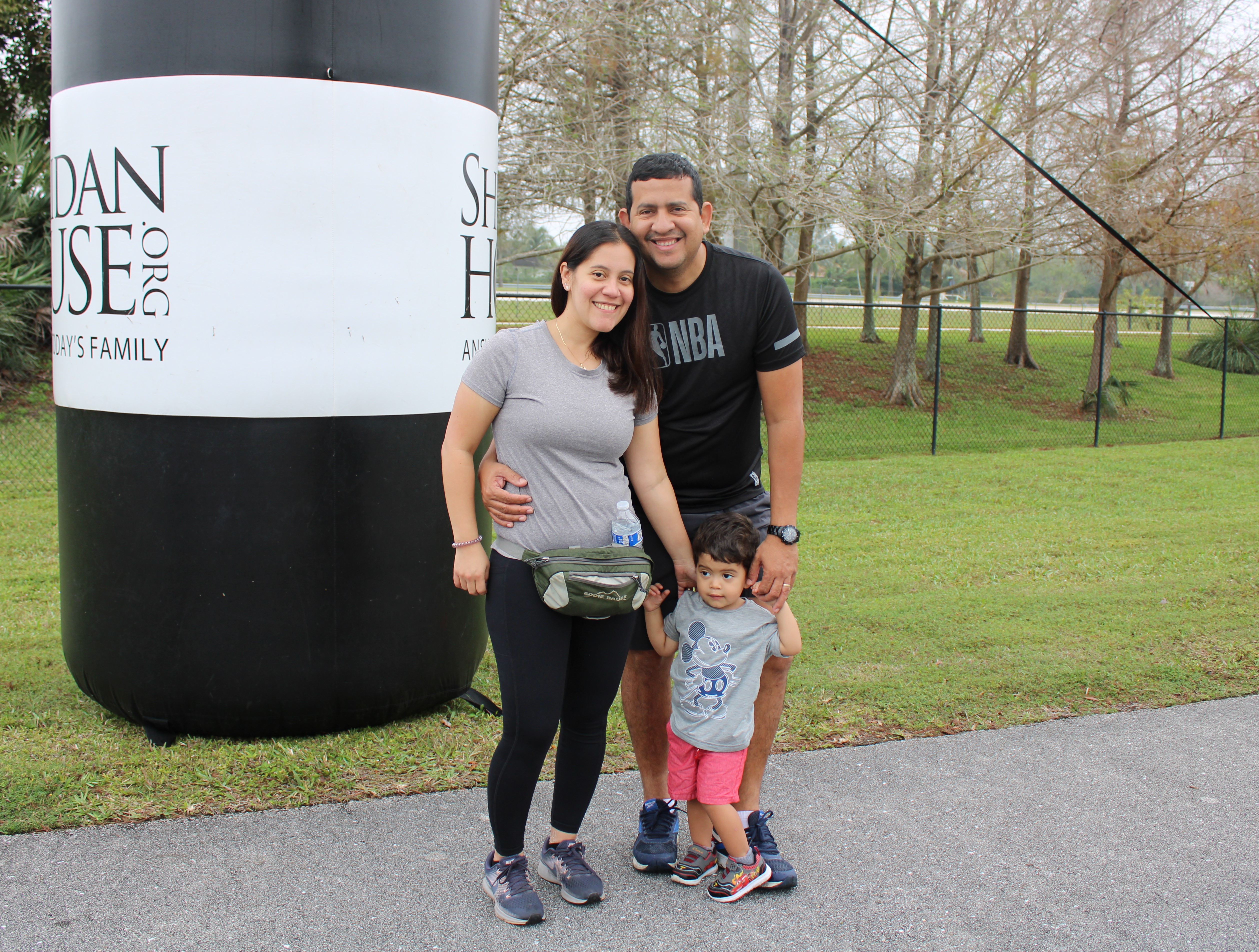 A smiling couple with a toddler stand in front of a black and white Sheridan House inflatable column.