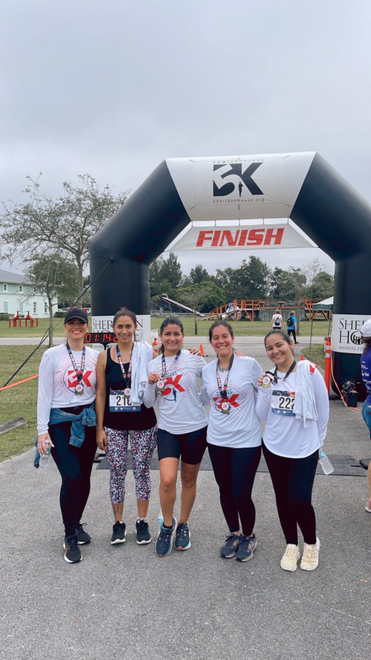 Five women hold up finisher medals in front of the FINISH arch, most in white long-sleeve shirts.