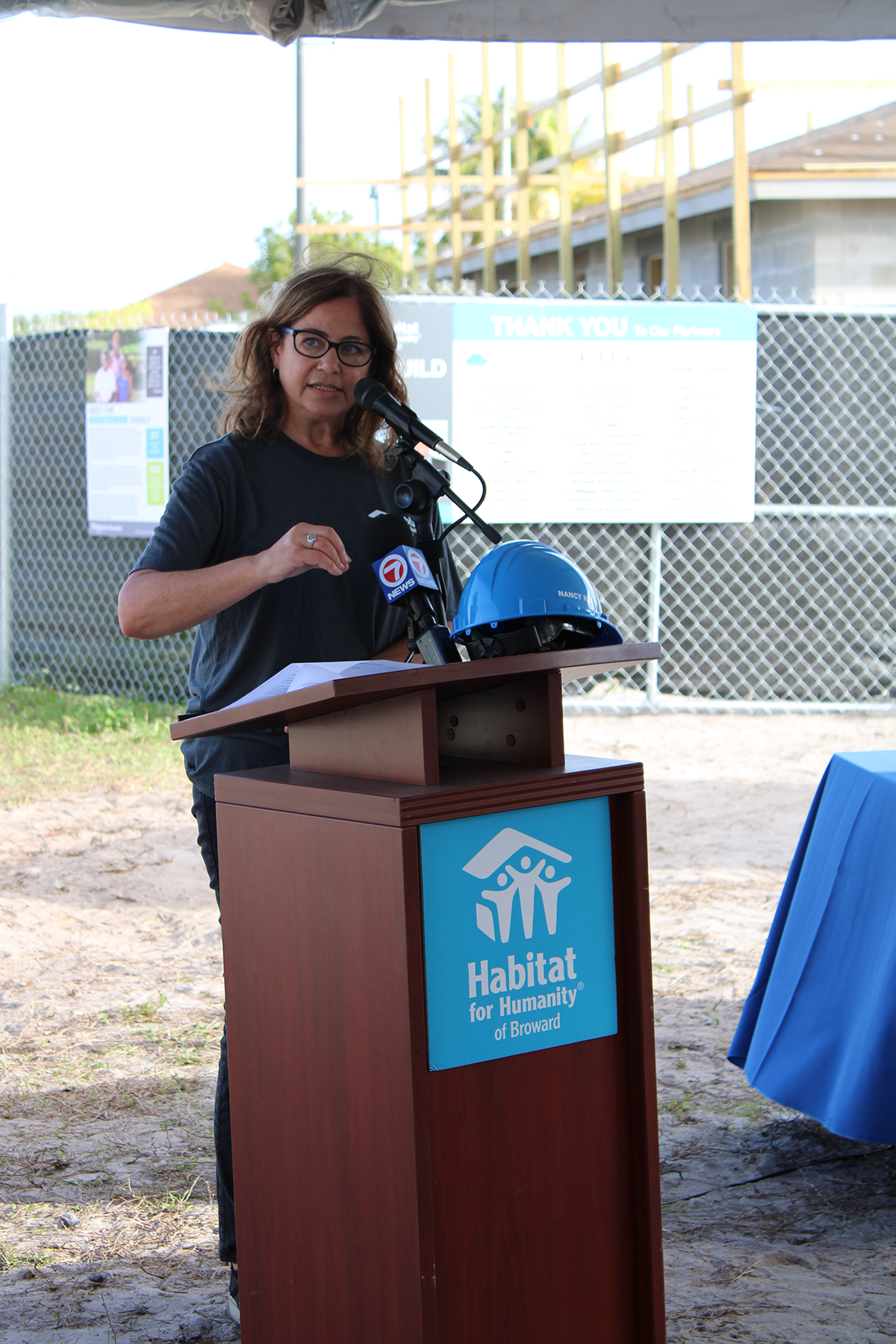 A woman in glasses speaks into microphones at a Habitat for Humanity of Broward podium, with a blue hard hat on it.