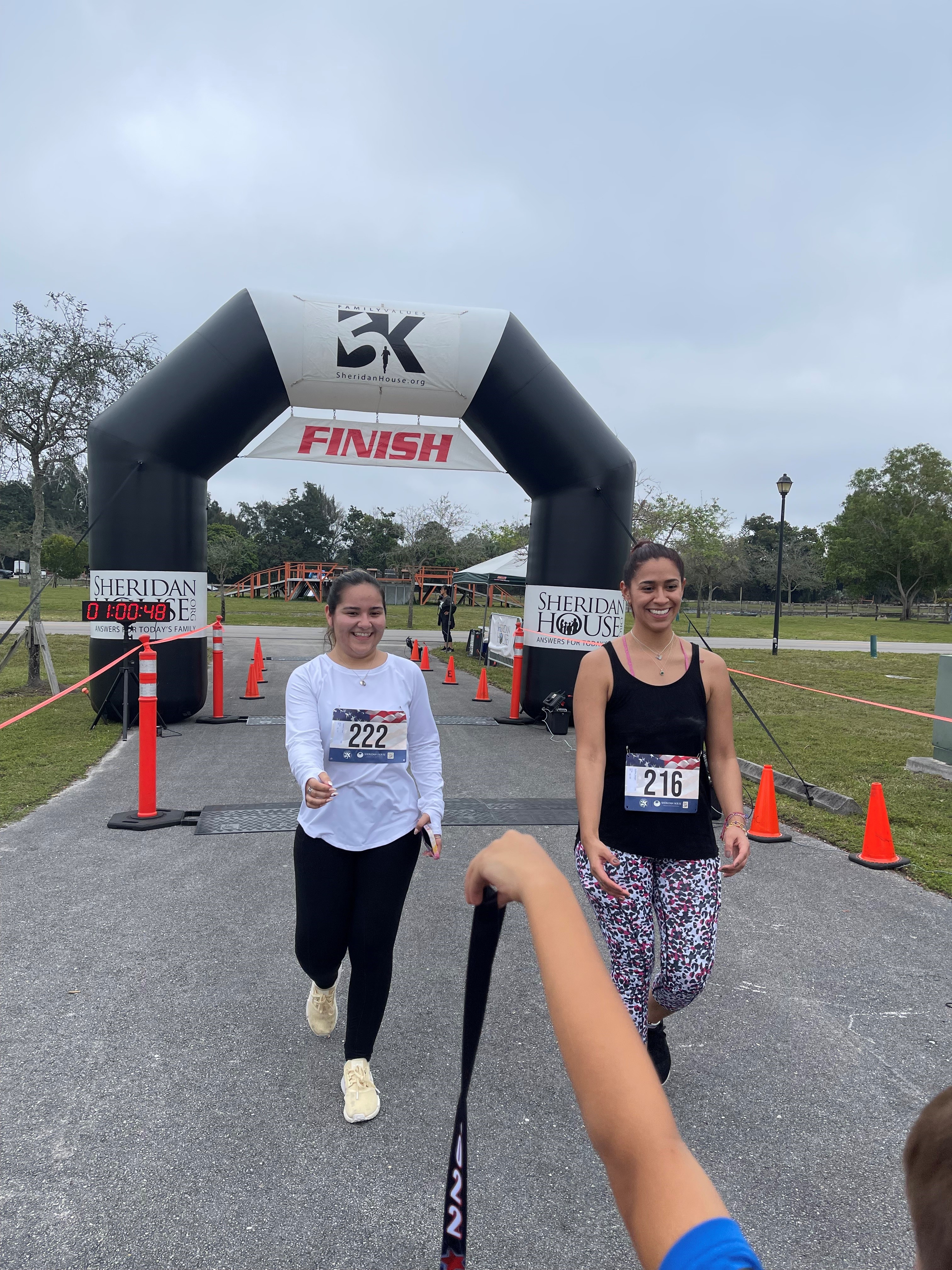 Two women smile as they cross under the FINISH arch, with a child's arm holding a medal ribbon in the foreground.