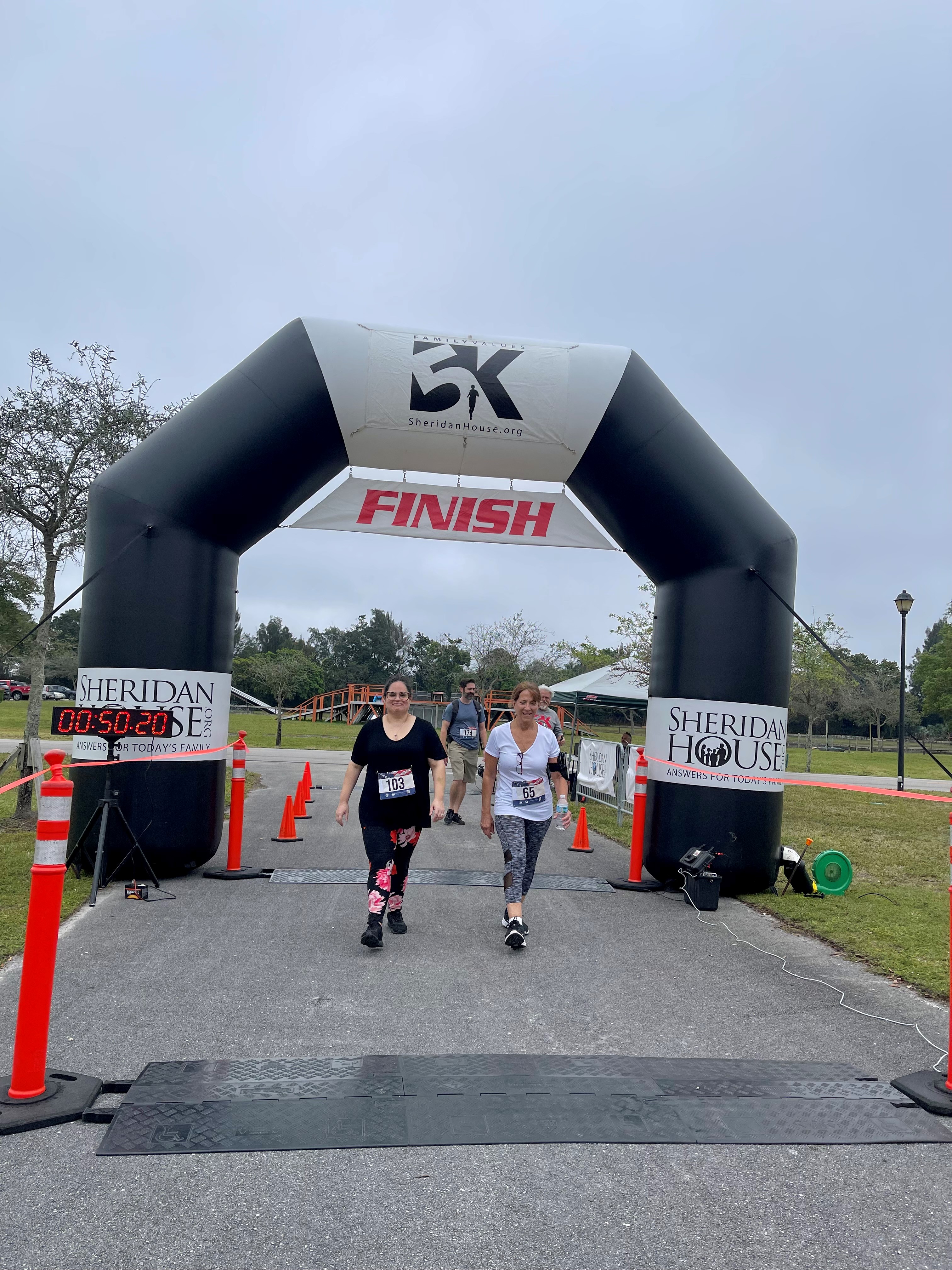 Two women in race bibs walk toward the camera under the FINISH arch beside a timer reading 00:50:20.