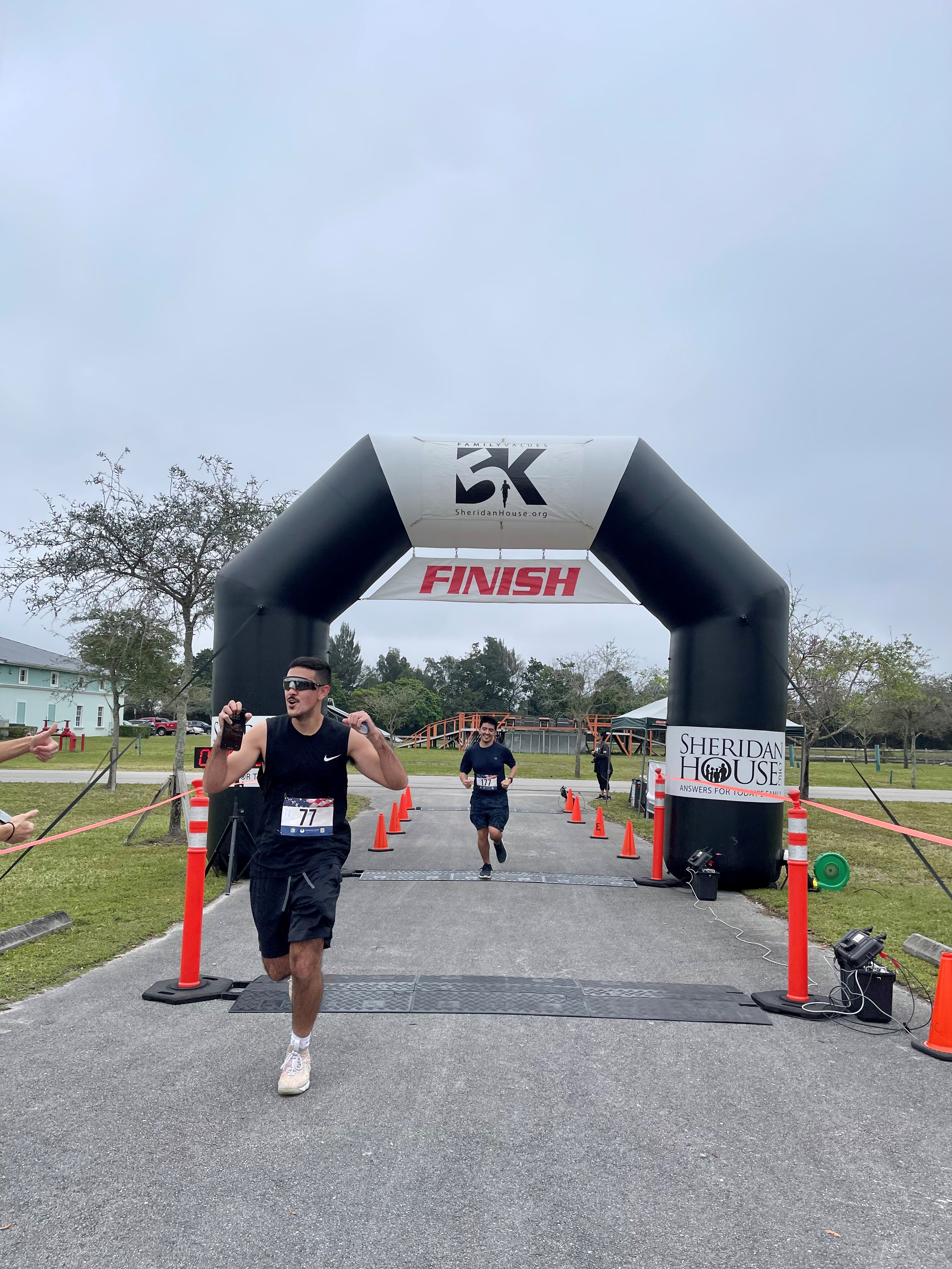 A man in sunglasses and a black tank top runs toward the FINISH arch, with another runner behind him.