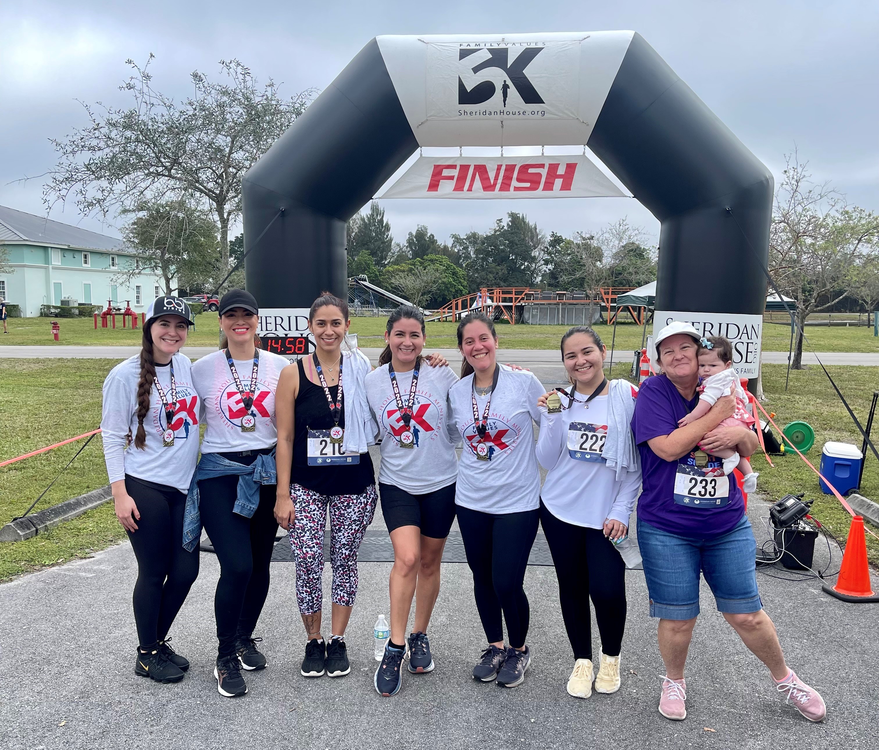 Seven participants pose in front of the FINISH arch, six wearing medals and one woman holding a baby.