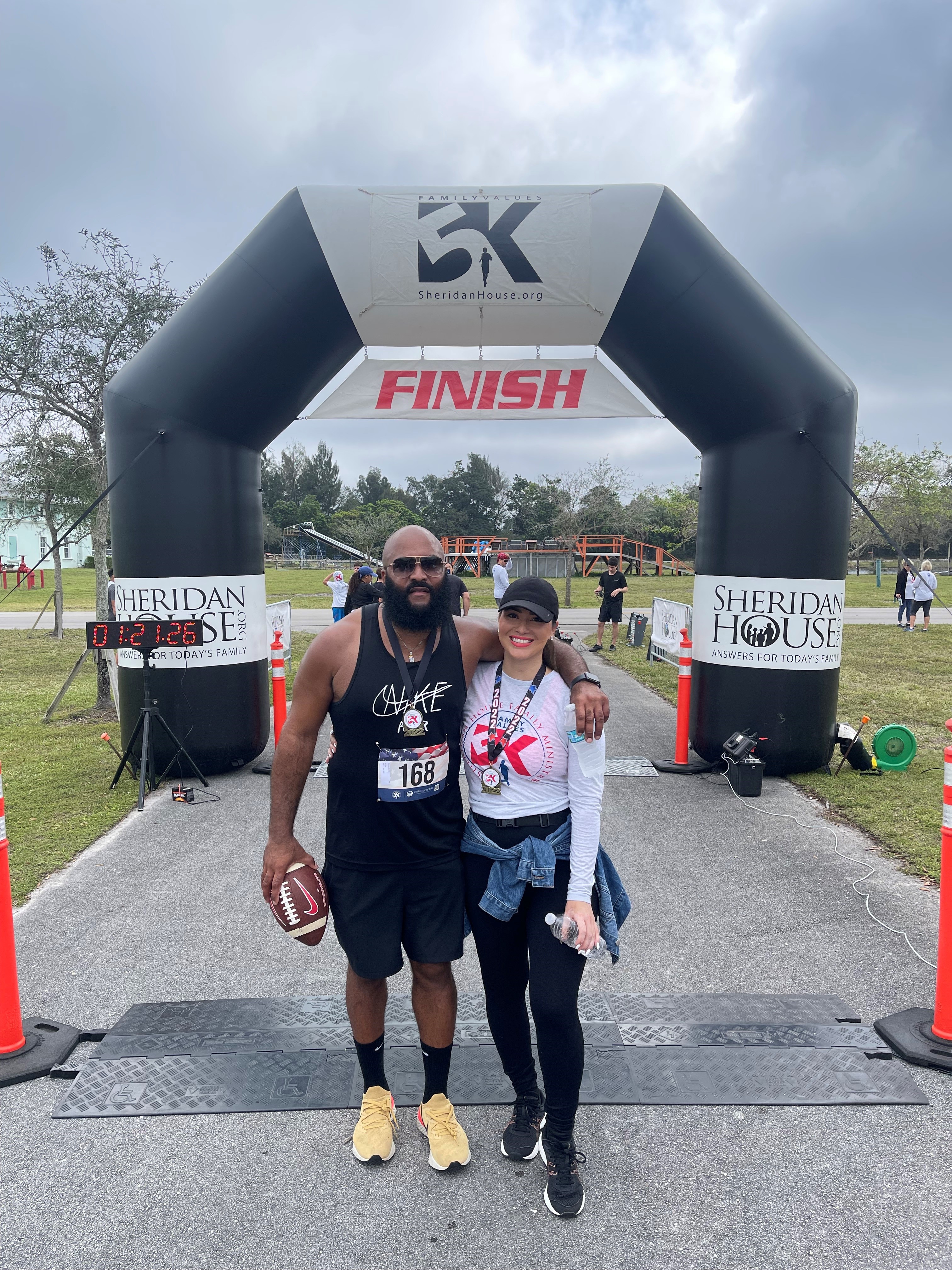 A bearded man holding a football and a woman in a cap pose arm in arm in front of the FINISH arch, both wearing medals.