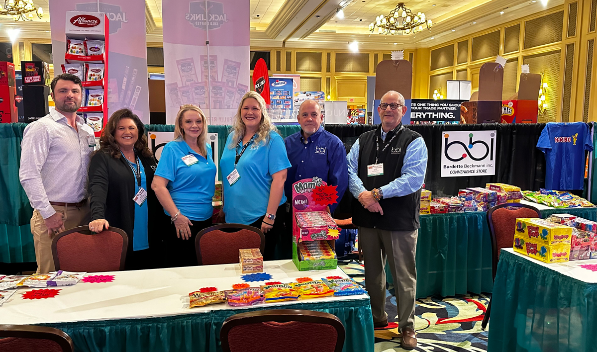 Six people, two in teal polos, stand behind a table with candy at a bbi booth in a chandeliered ballroom.
