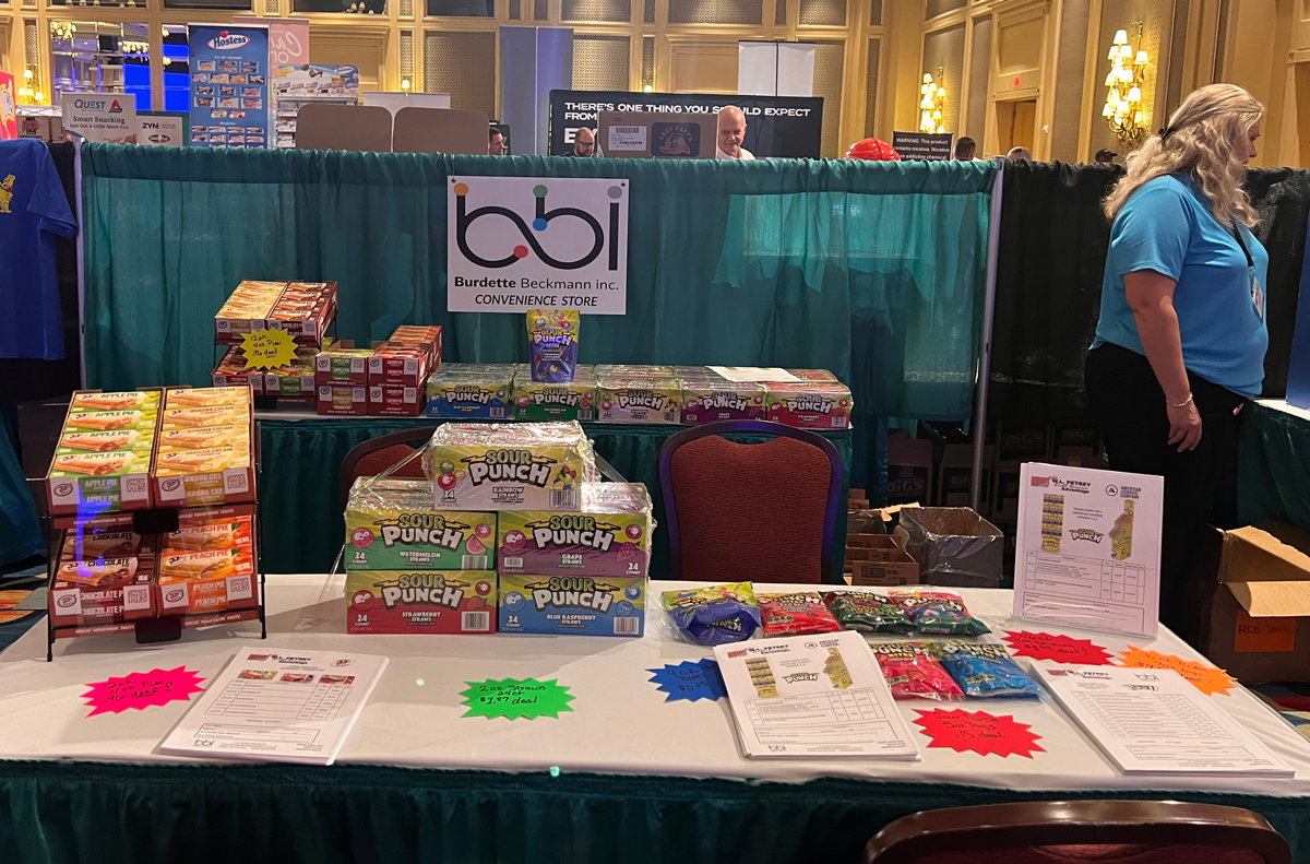 Table with stacked Sour Punch cases, price sheets and a candy rack, with a bbi sign on teal drapes behind.