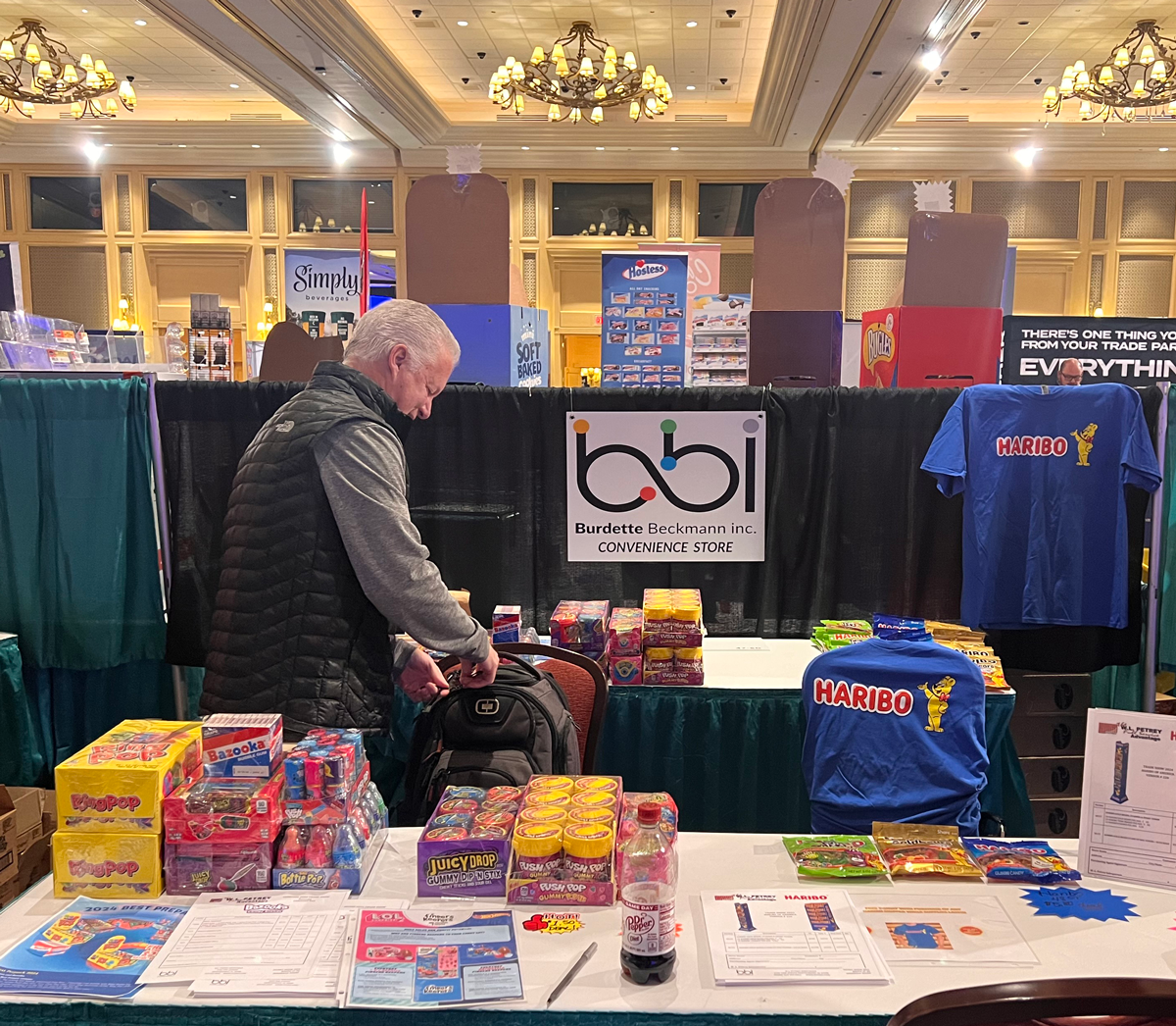 Man in a black vest looks into a backpack at a table with Haribo blue shirts, Dr Pepper and candy boxes.