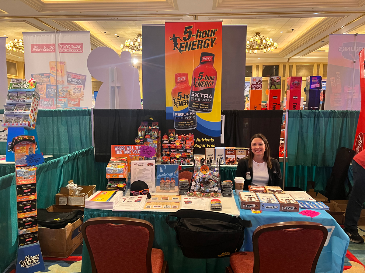 Woman seated at a booth behind a 5-hour ENERGY banner and display, with a Juicefuls rack at left.