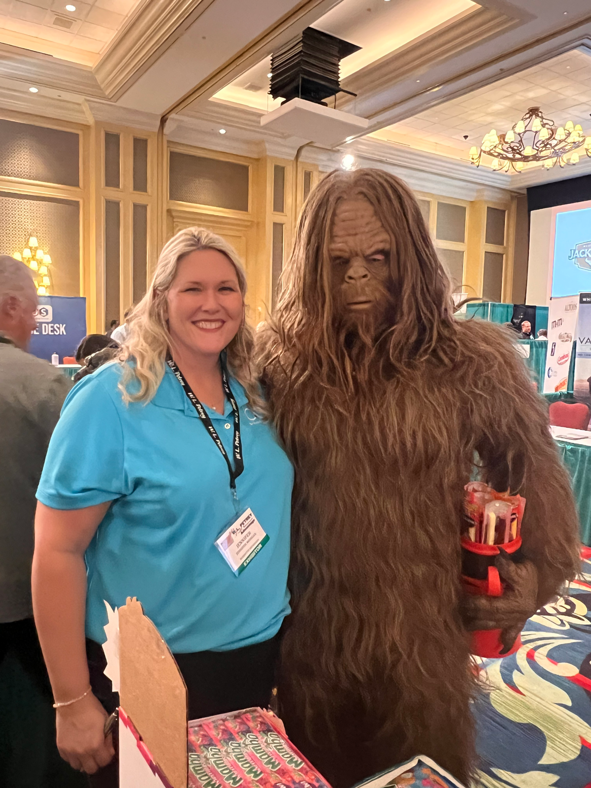 Smiling woman in a teal polo beside a person in a Bigfoot costume holding a red container.