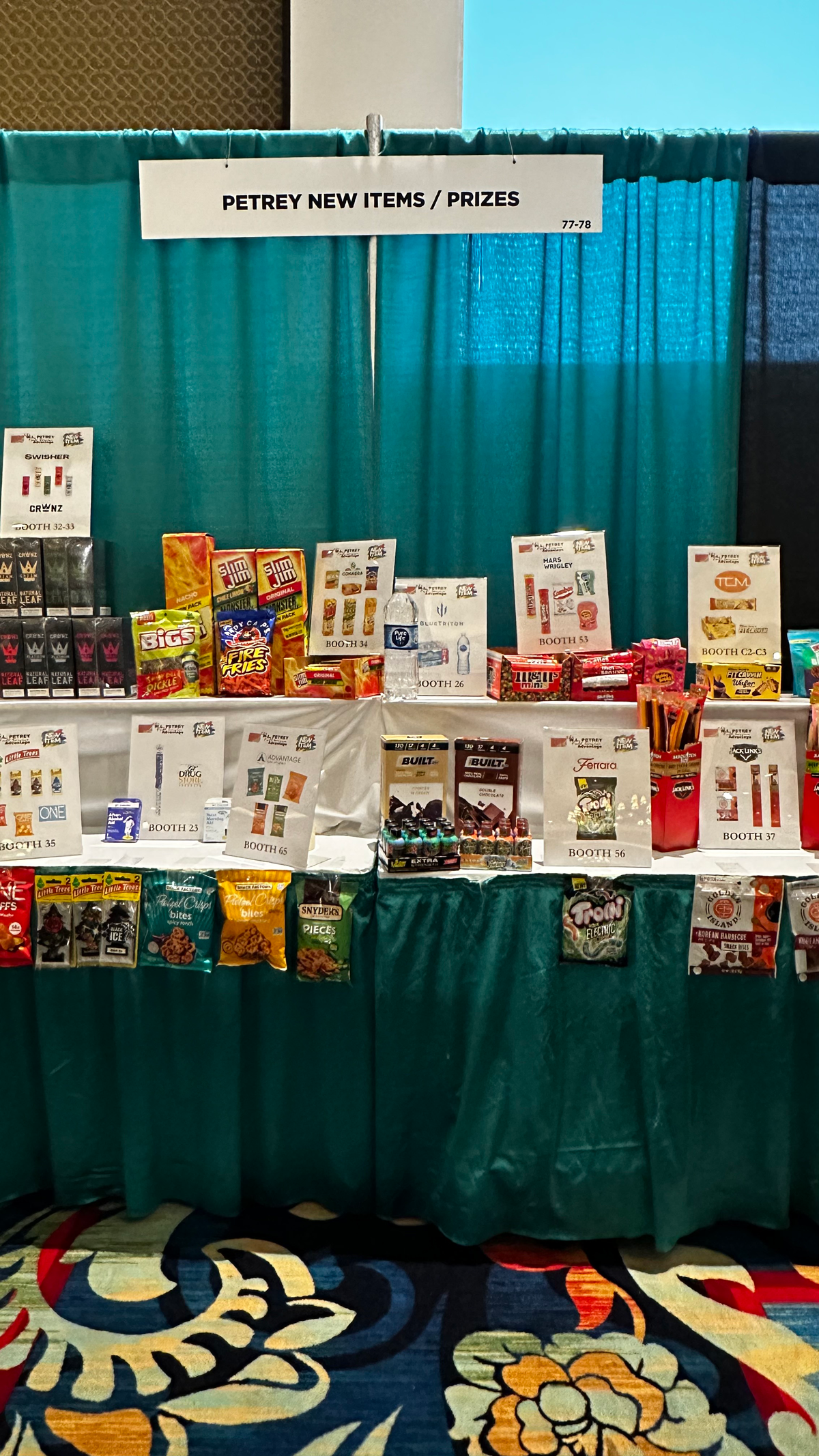 Table display under a sign reading Petrey New Items and Prizes, with snack packages and booth number cards.