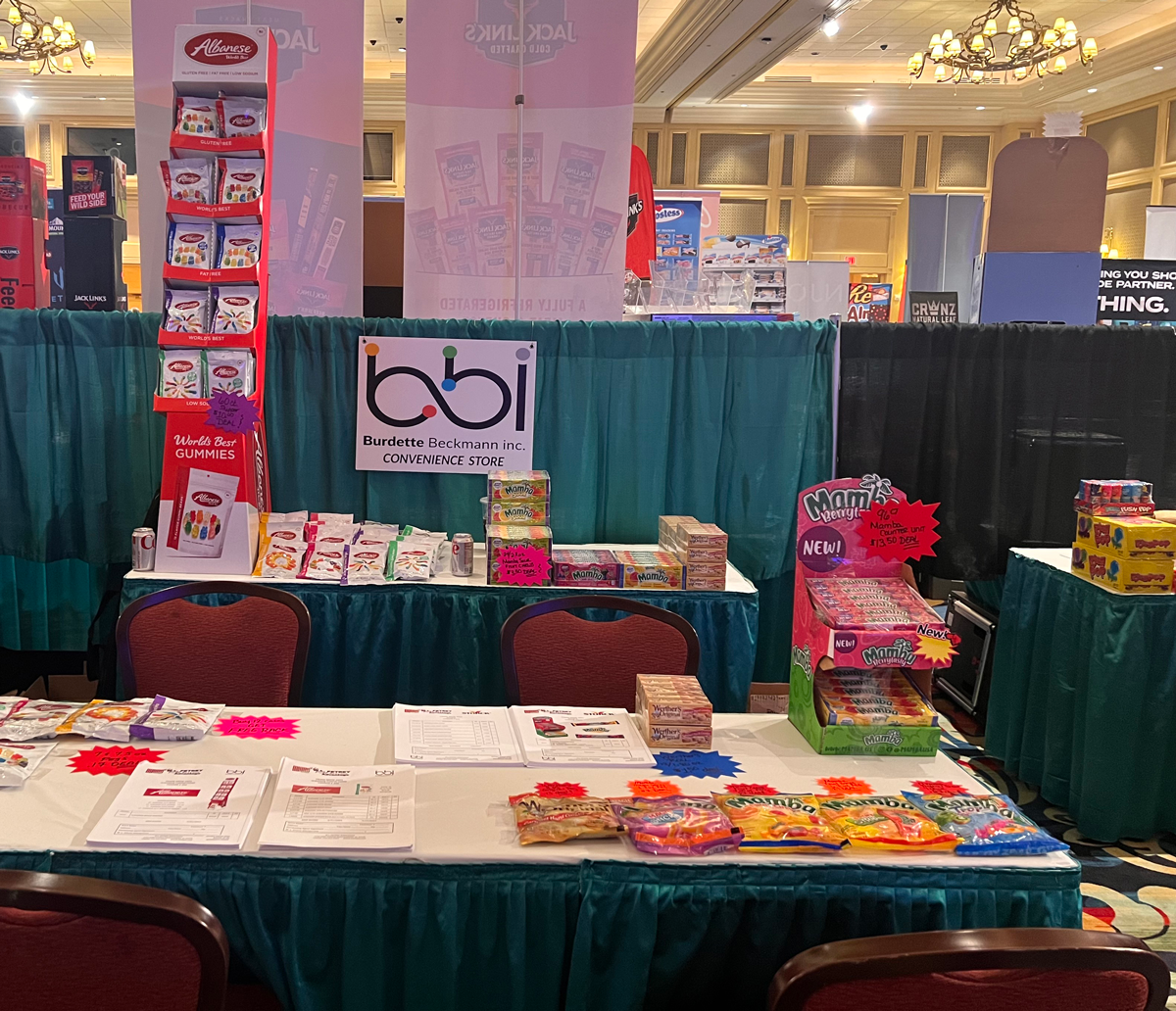 Unstaffed booth with a bbi Convenience Store sign, an Albanese gummy rack, Mamba chews and price sheets on a table.