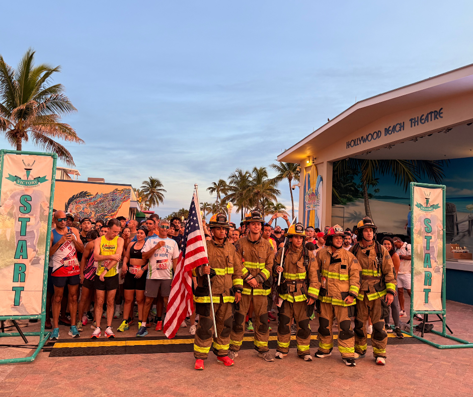 Five firefighters in full gear, one holding a U.S. flag, stand at the Start line before a crowd of runners.