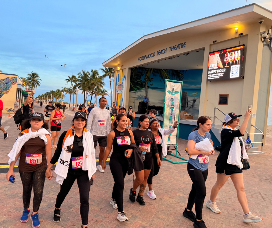 Walkers in race bibs, including a woman in a bbi shirt, stroll past the Hollywood Beach Theatre and Start sign.