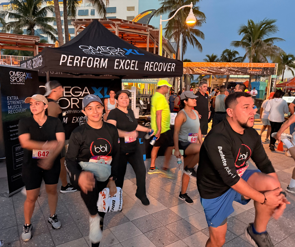 Participants in bbi shirts and bibs stretch beneath an Omega Sport XL tent on the beachfront promenade.