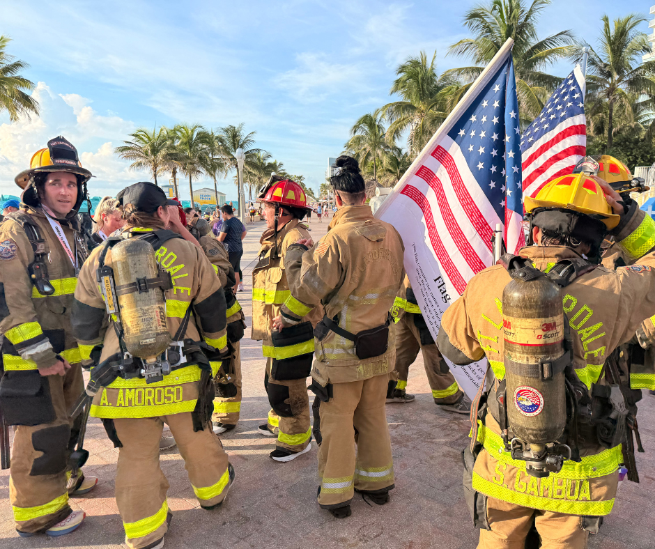 Firefighters in tan gear with air tanks stand near large American flags on a palm-lined walkway.