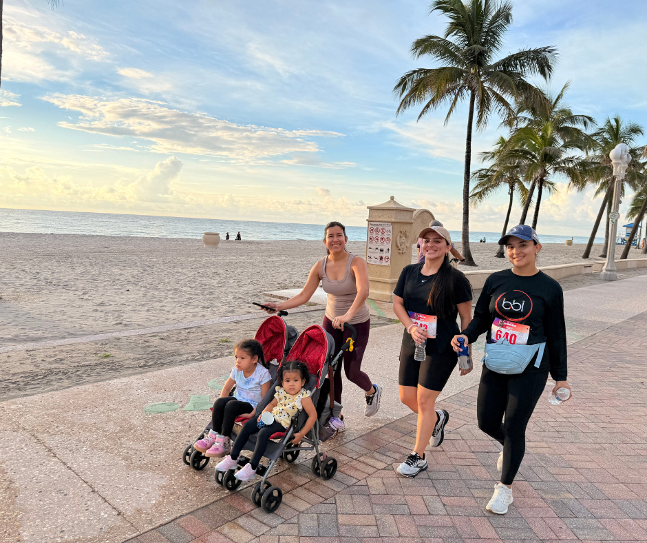 Woman pushes a double stroller with two toddlers on the beach boardwalk beside two women, one in a bbi shirt.
