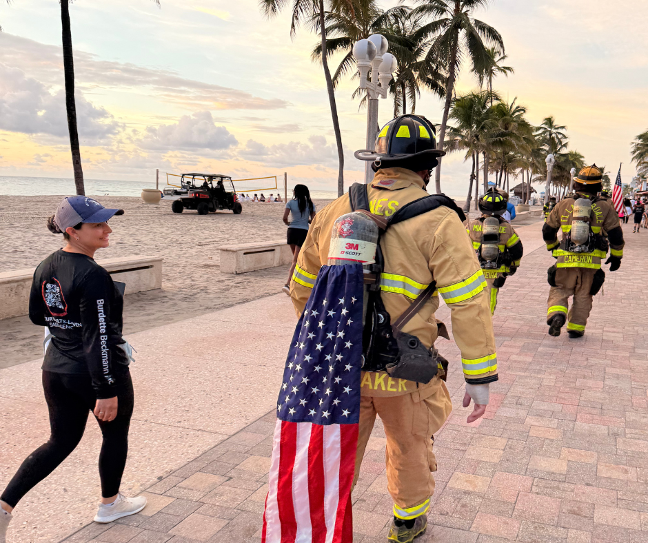 Firefighter in full gear carries an American flag along the beach boardwalk past a woman in a bbi long-sleeve shirt.