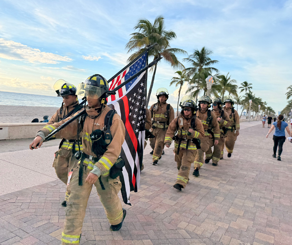Line of firefighters in helmets march along the beachfront carrying a thin blue line American flag.